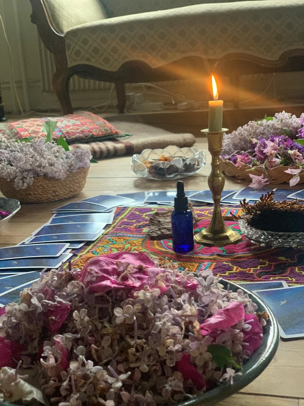 A cozy scene of a table set for a tarot or oracle card reading, with a lit candle, flowers, a blue dropper bottle, and various cards arranged on a colorful cloth, in a softly lit room.