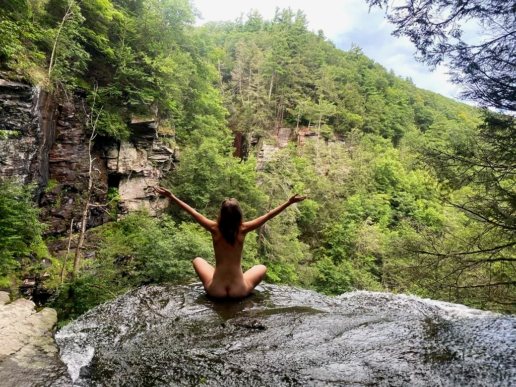 A woman sitting naked at the edge of a natural rock pool overlooking a forested canyon, with arms outstretched.