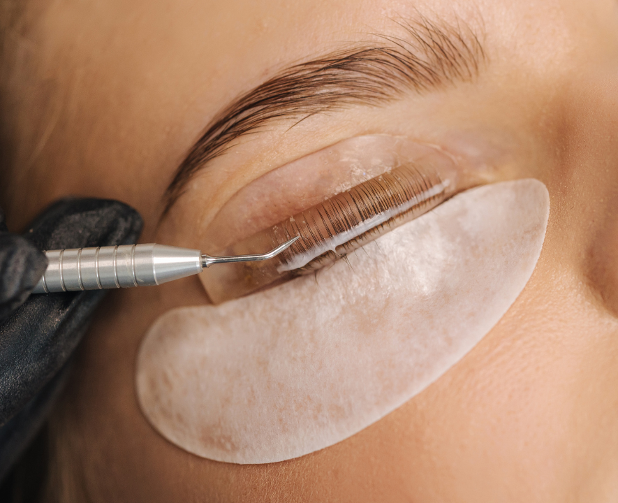 A person getting eyelash extensions applied by a Korean technic using a precision tool. The person's eye is closed with an under-eye patch and a protective adhesive pad.
