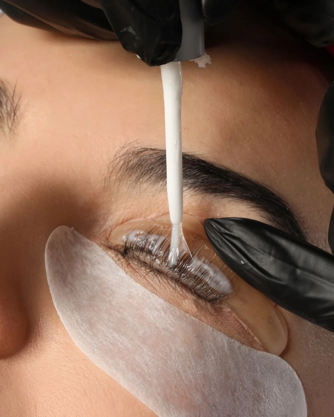 A person receiving an eyelash extension or eyelash treatment with a technician applying a liquid adhesive or serum to the eyelashes. The person's eye is closed, with a protective pad placed under the eye, and the technician wearing black gloves.