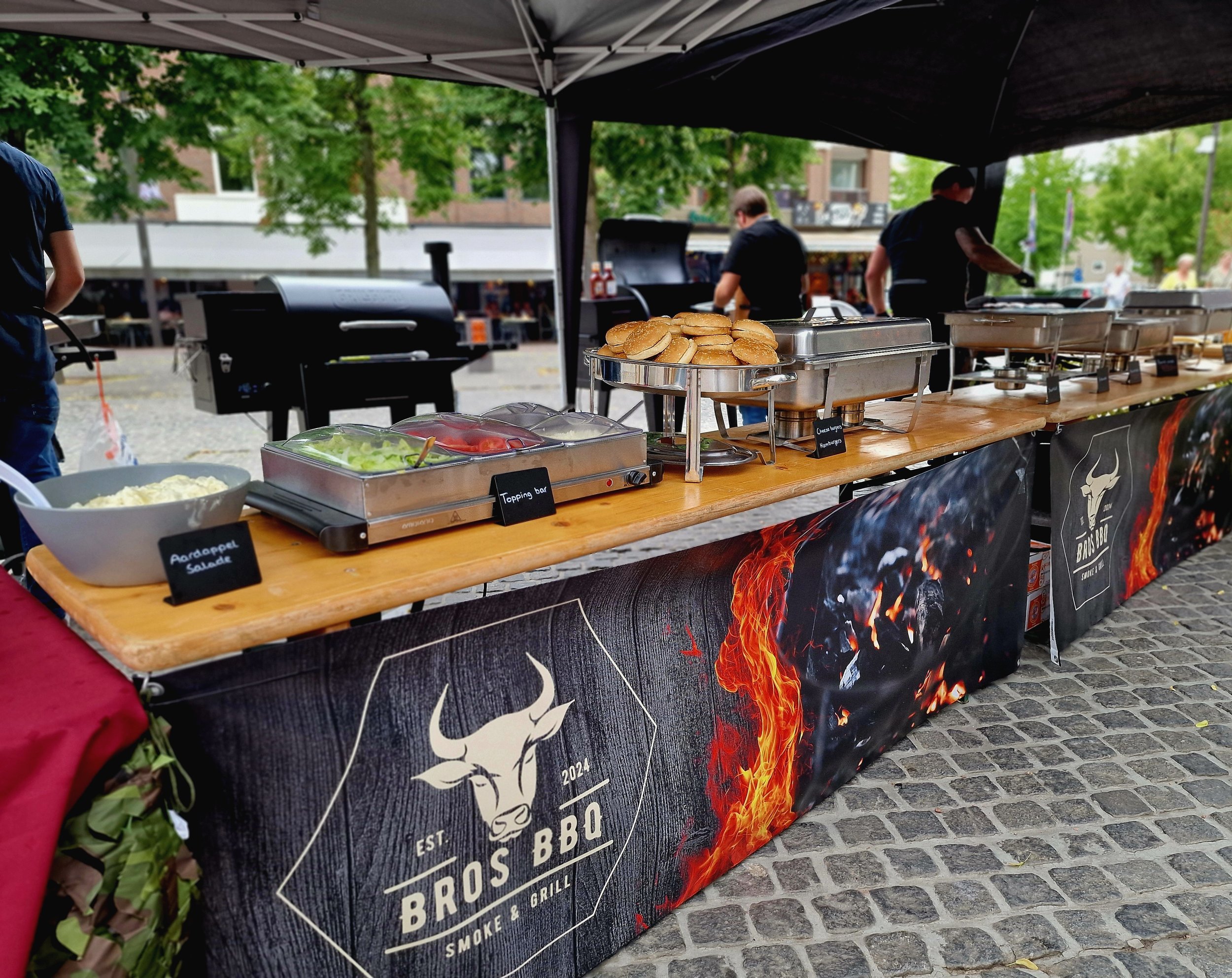 Een food truck of stand met BBQ en sandwich vlees en diverse toppings, op een straat met bomen en mensen op de achtergrond.