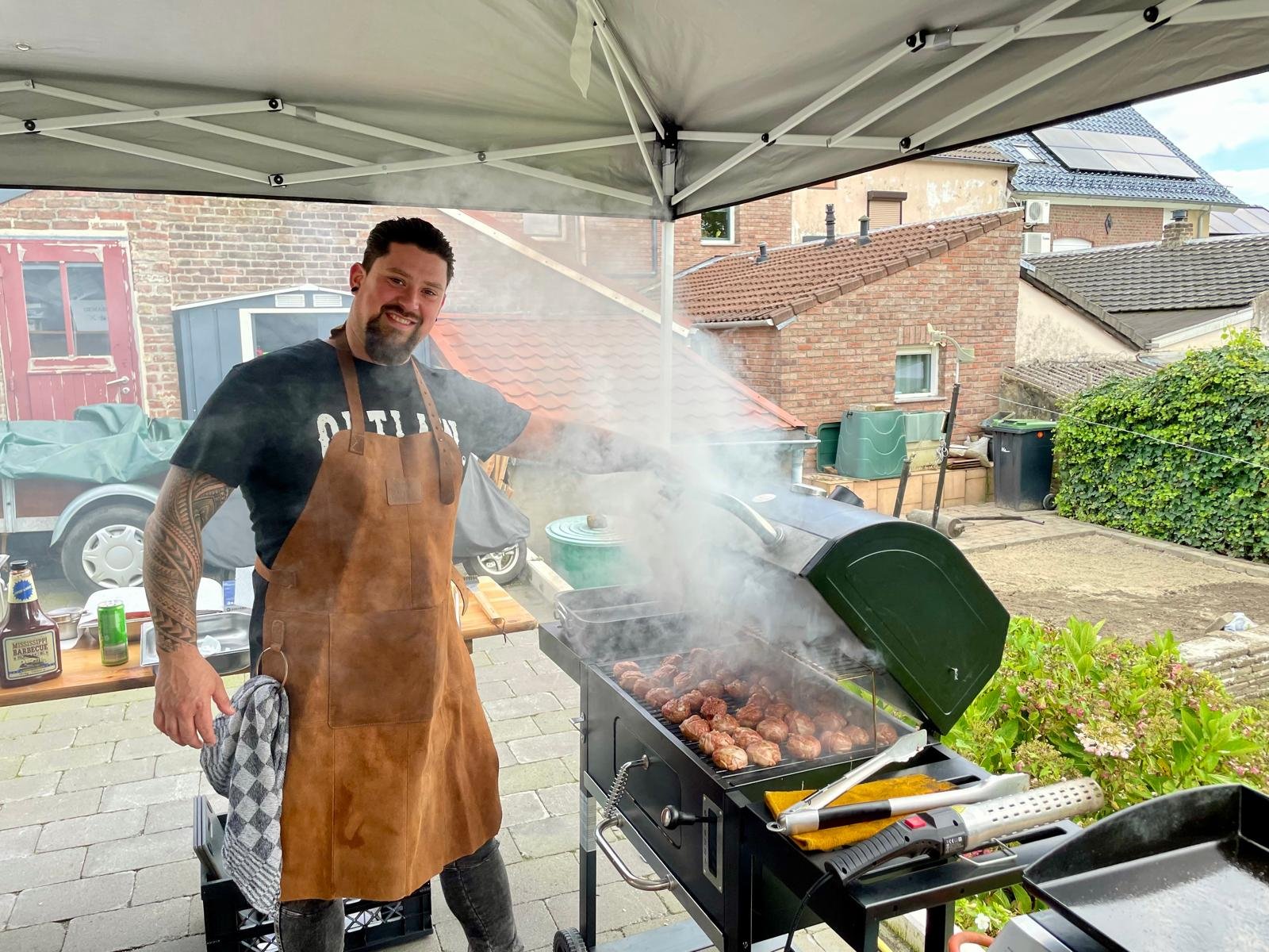 Man die gegrilde vlees en gehaktballetjes bereidt op een barbecue onder een tent op een patio met huisdaken op de achtergrond.