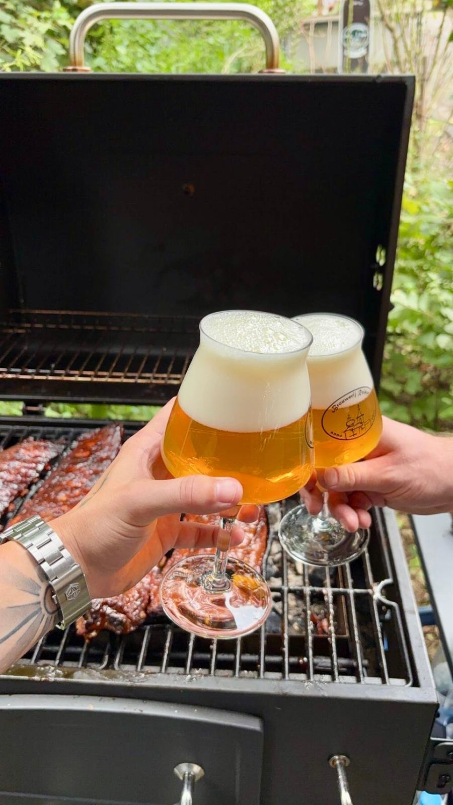 Drie mensen heffen glazen met bier in de handen boven een barbecue met vlees, in een tuin met groene planten.