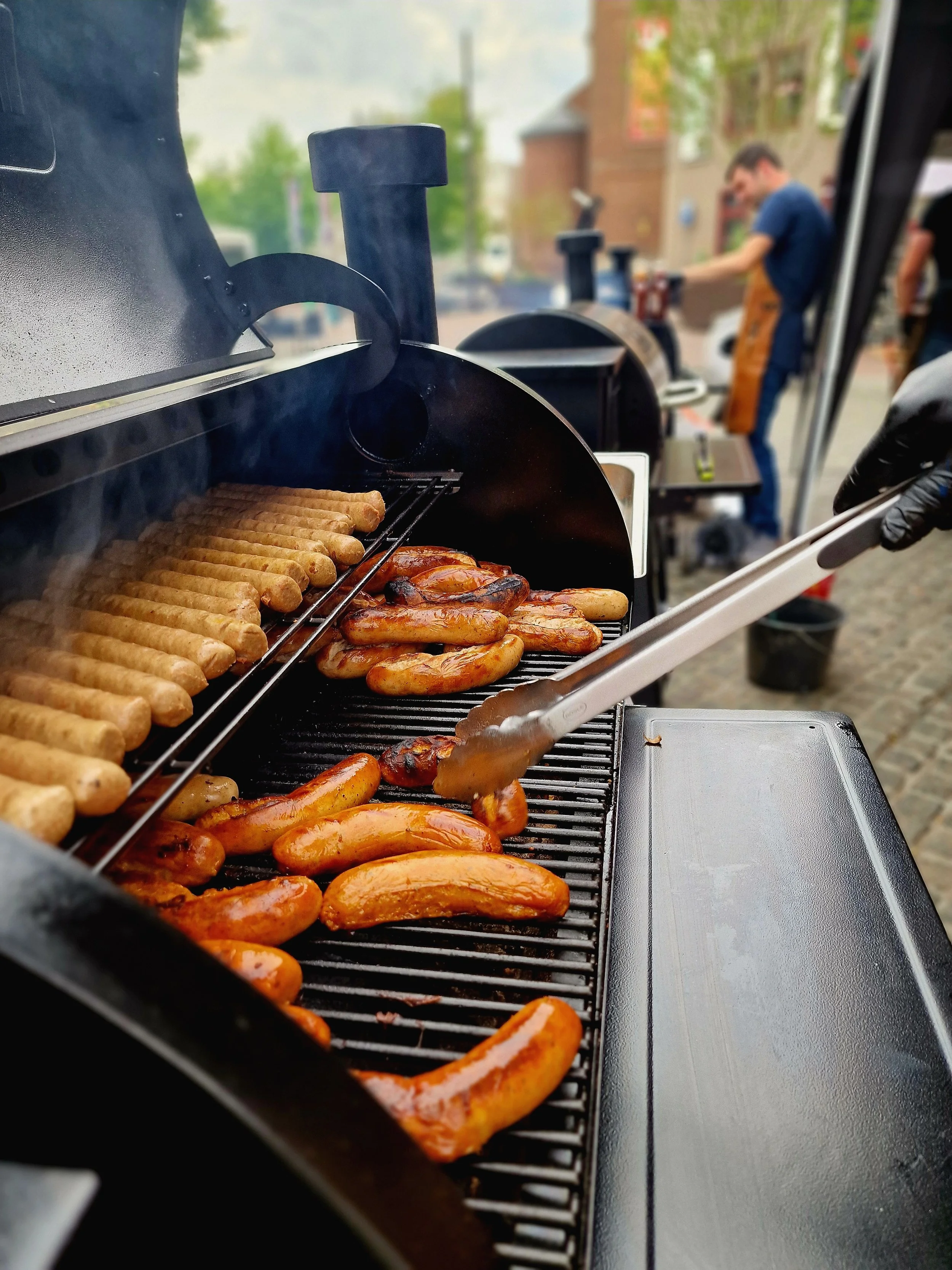 Barbecue met verschillende soorten vlees en worst, personen die grillen op een buitengrill, in een stadsscene met gebouwen op de achtergrond.