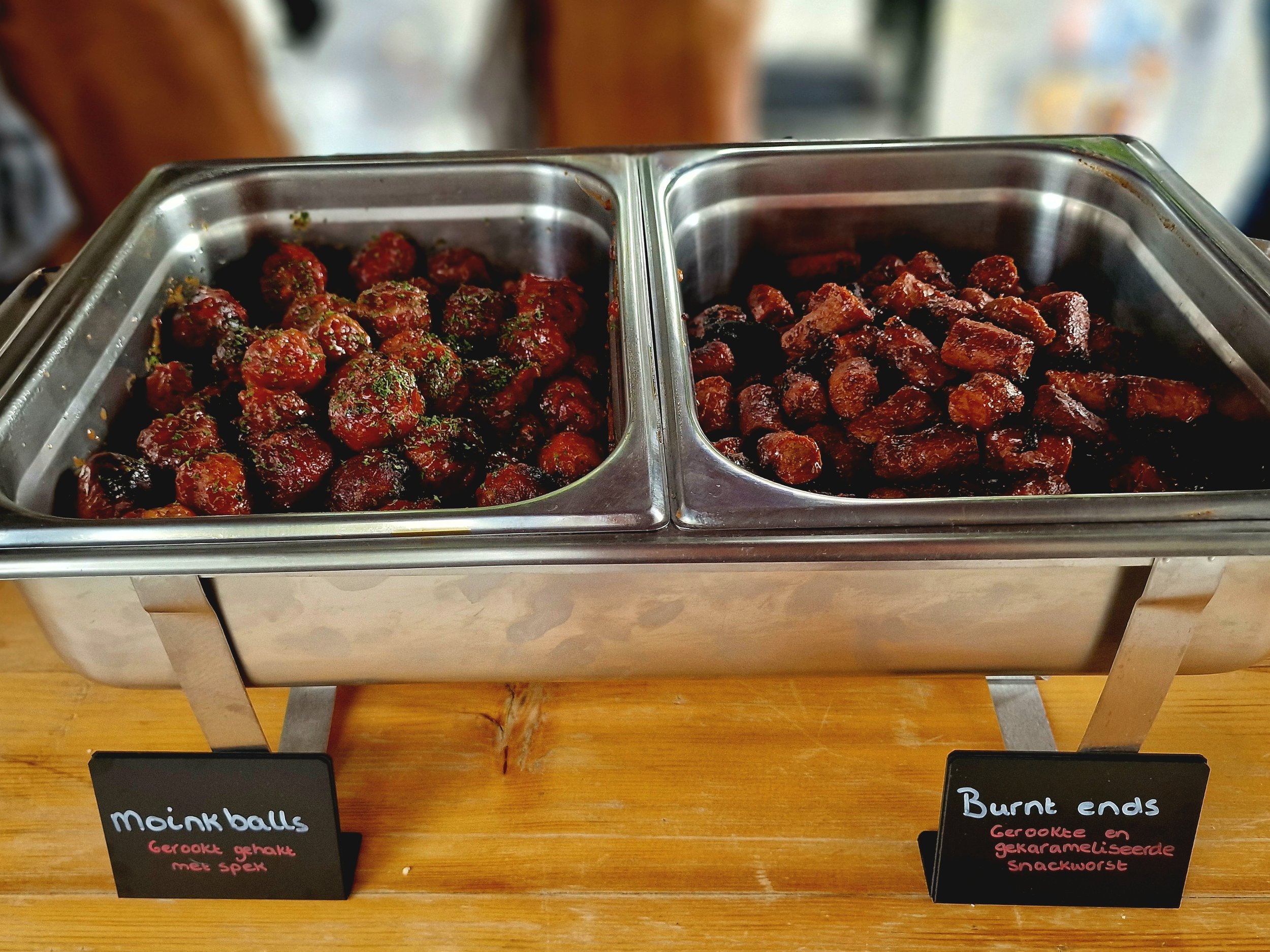 Two metal trays containing smoked meatballs and burnt ends on a wooden surface, with small black signs labeled in Dutch.