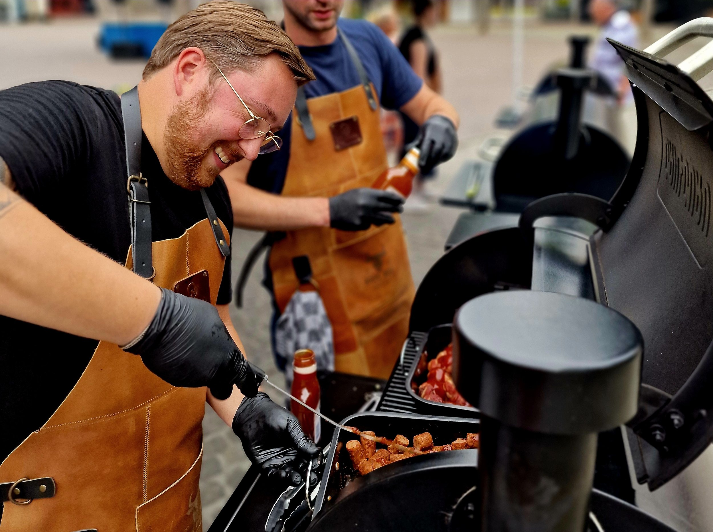 Mensen die barbecueën in een buitenomgeving, met één persoon die vlees en worstjes op de grill legt. Ze dragen all weather kleren en leren of rubberen handschoenen.