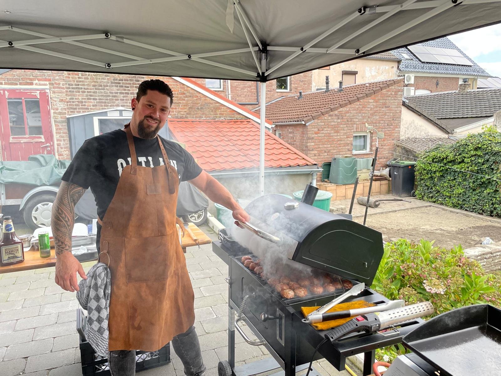 Man met tatoeages en een bruin leren schort grillt vlees op een barbecue onder een tentdoek in een tuin met huizen op de achtergrond.