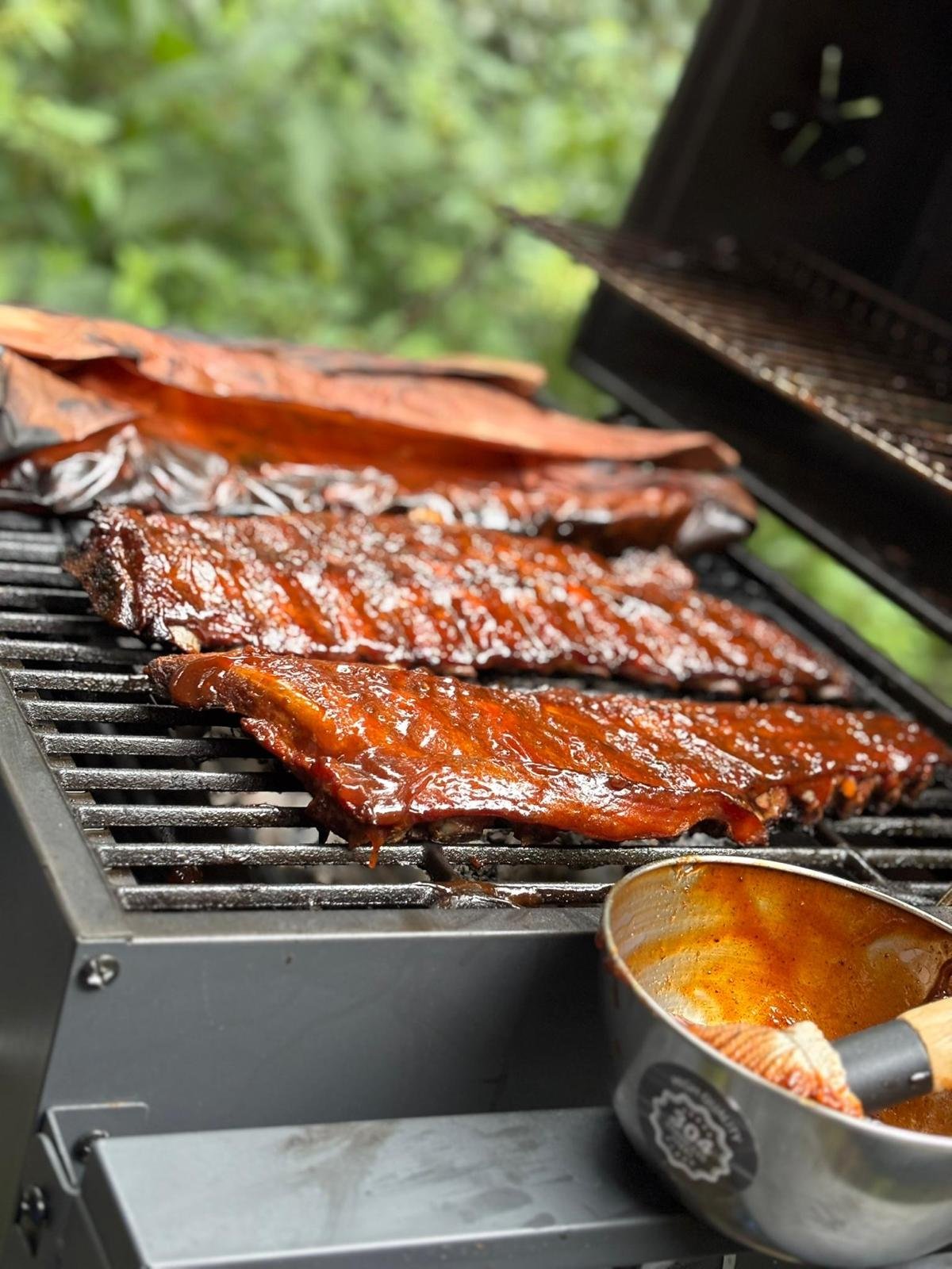 Barbecue met gerookte spareribs en vlees in een saus, met een bakje saus naast de grill, in een groene omgeving.