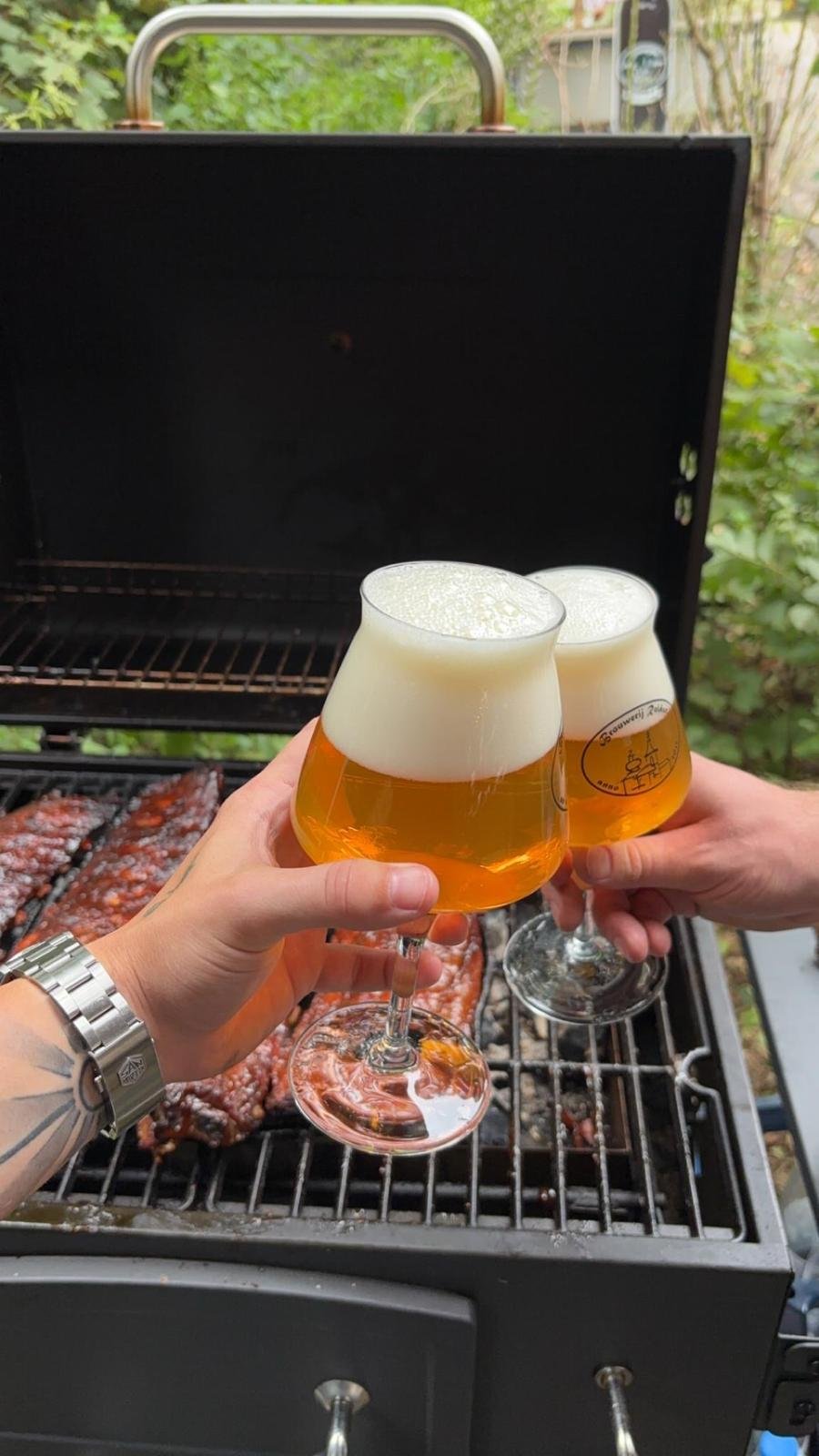 Twee mensen proosten met glazen gekoeld bier boven een barbecue met gerookt vlees in de tuin.