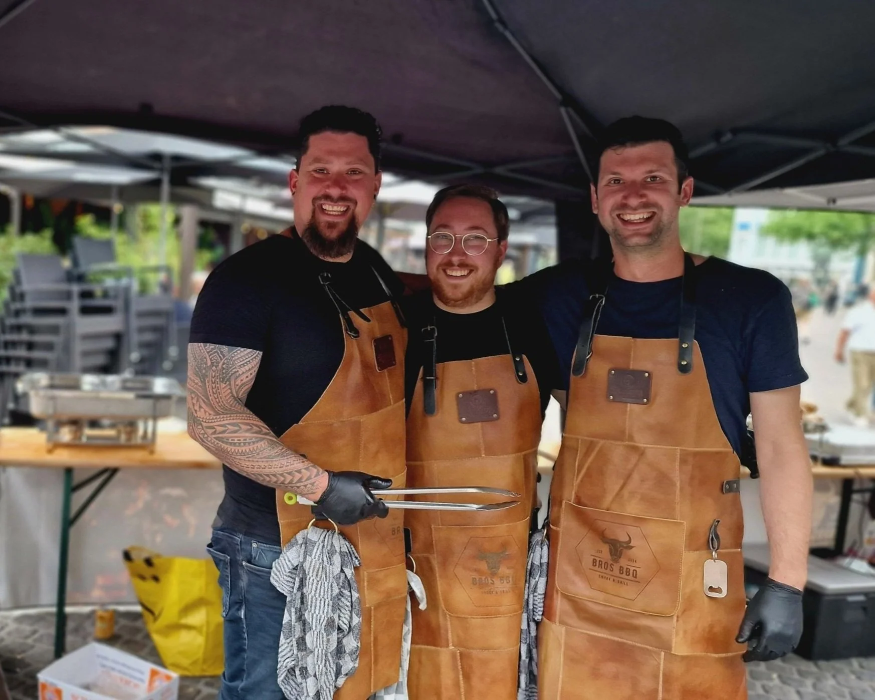 Drie mannen in zwart T-shirt en babyribben, die een leren schort dragen, staan onder een tent en glimlachen naar de camera. Ze lijken klaar voor een barbecue.