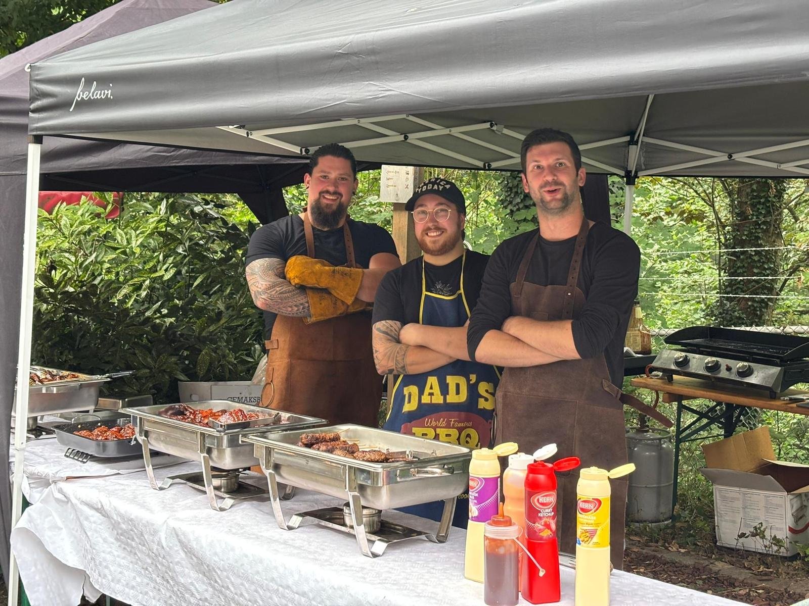 Drie mannen staan achter een barbecuetafel onder een tent, met diverse sauzen en vlees op het buffet. Ze dragen aprons en lachen naar de camera in een groene, open lucht omgeving.