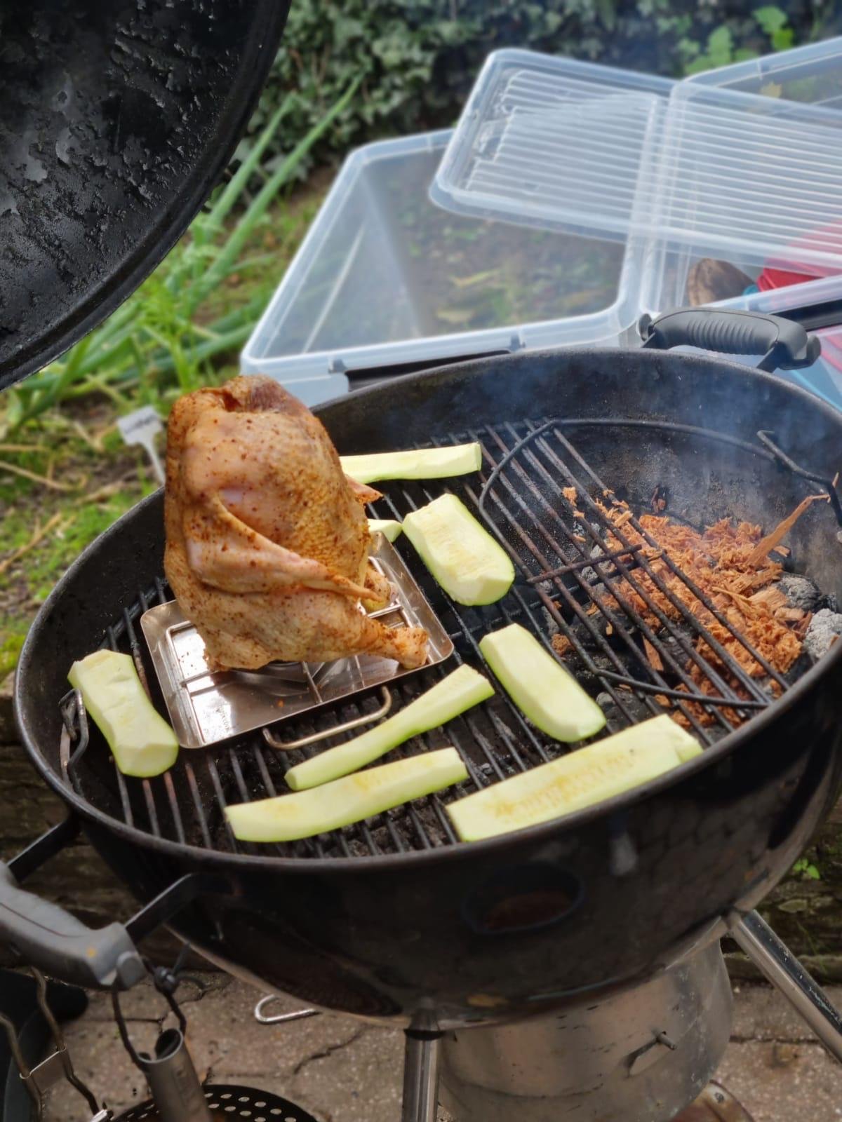Een barbecue met een geroosterde kip, komkommerstukjes en wat vlees, buiten met tuin en tuinset in de achtergrond.
