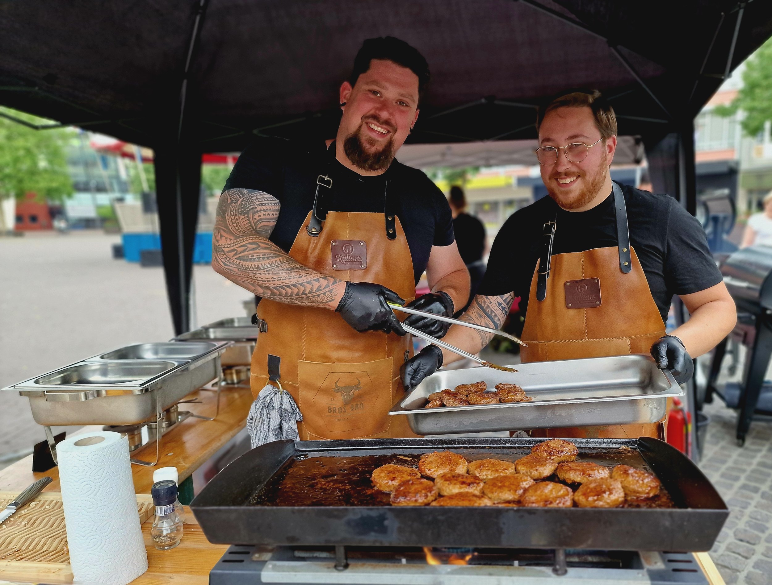 Twee mannen die barbecuen, vlees op de grill en in bakplaat, onder een zwart tentdoek, op een buitenevenement.