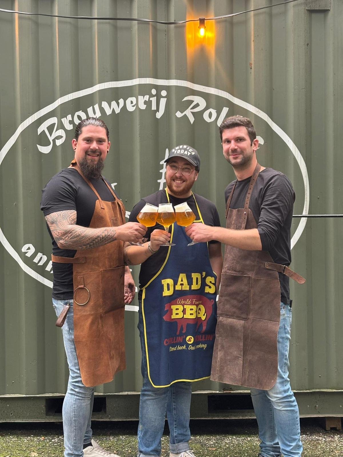 Drie mannen in leren schorten proosten met glazen bier voor een groene container met witte tekst 'Brouwerij Rol'.
