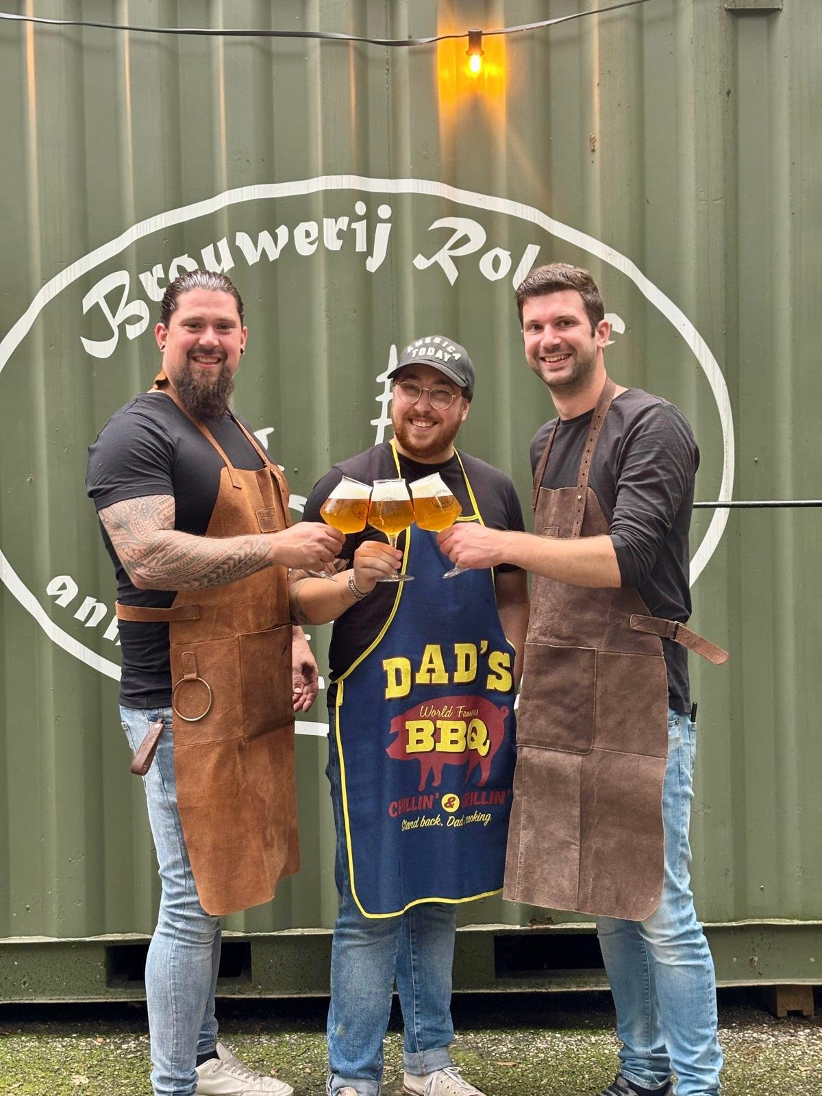 Driemen in leren schorten proosten met bier voor een groene muur met witte tekst, waarschijnlijk bij een brouwerij of barbecue evenement.