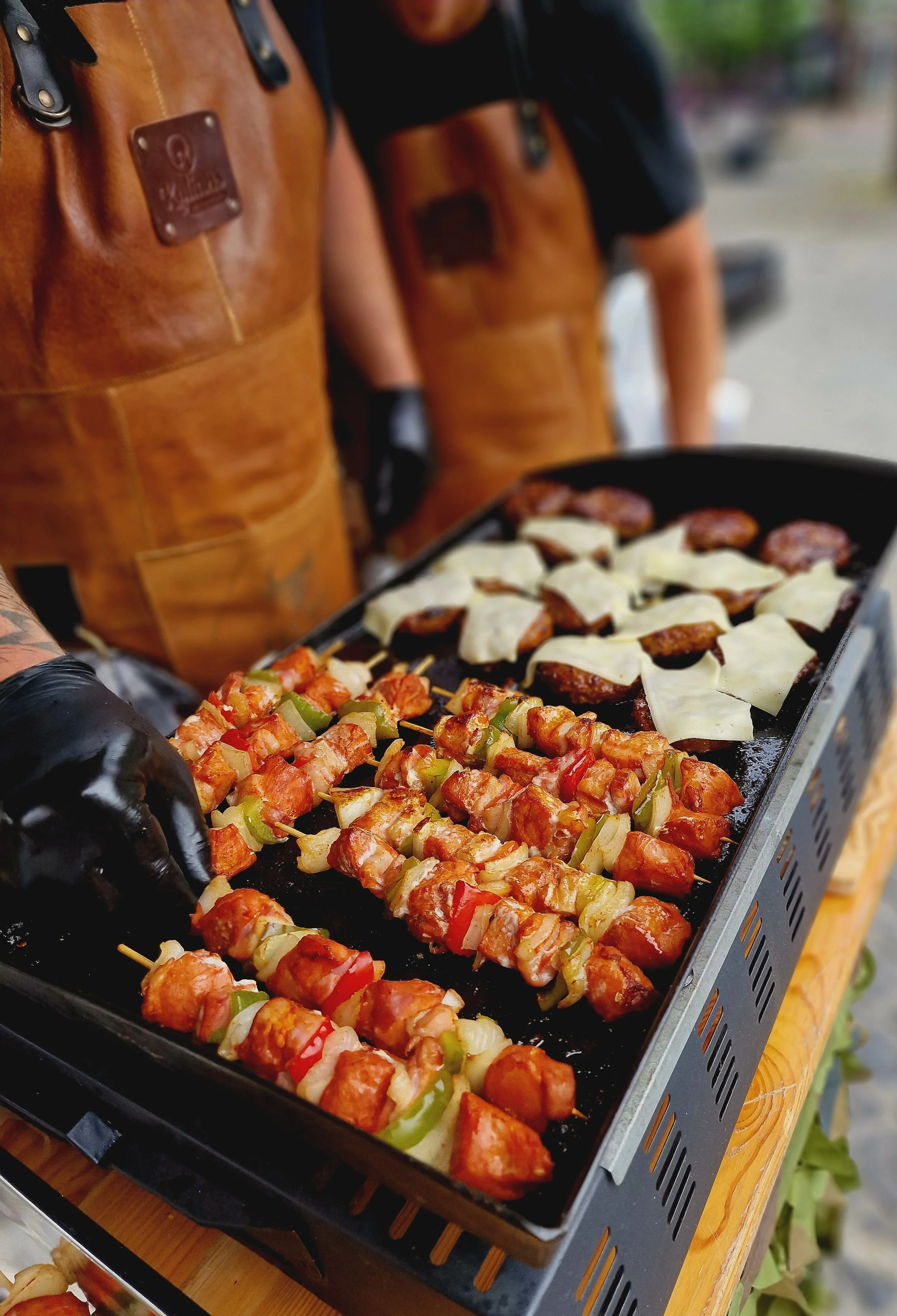 Barbecue met kipspiesjes met groenten en hamburgers met kaas op een grill, man in leren schort en handschoenen, buitenmarkt of evenement.
