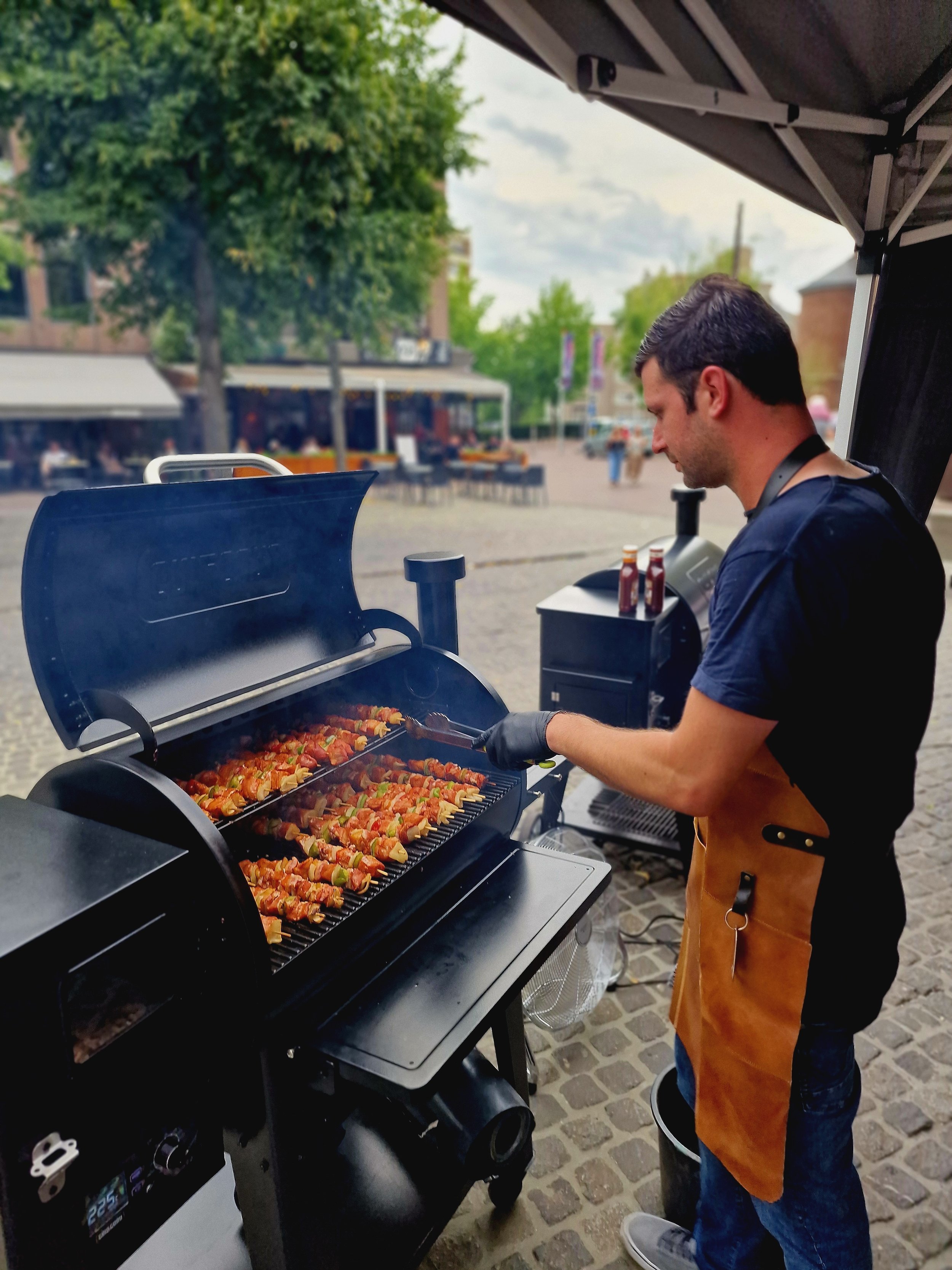 Man die barbecuet met spek en groenten op een grill, buiten op een plein onder een tent.