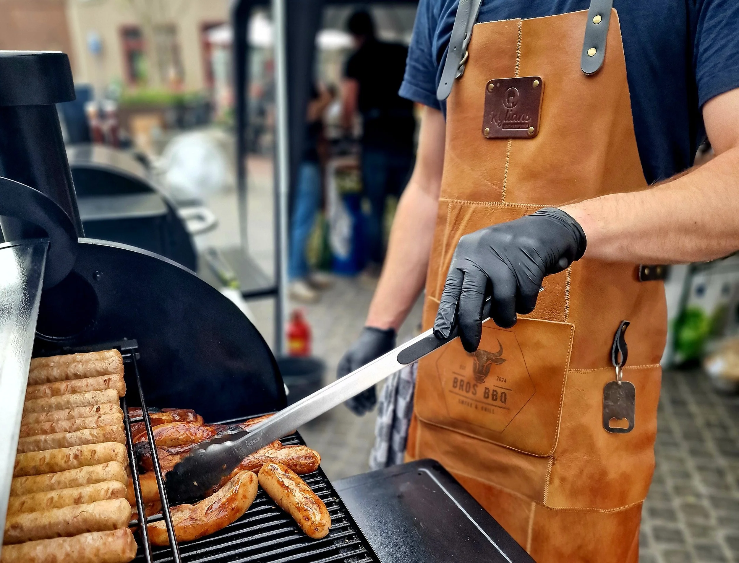 Persoon die barbecue vlees en worstjes op een grill kookt, gekleed in een bruine leren schort en zwarte handschoenen.
