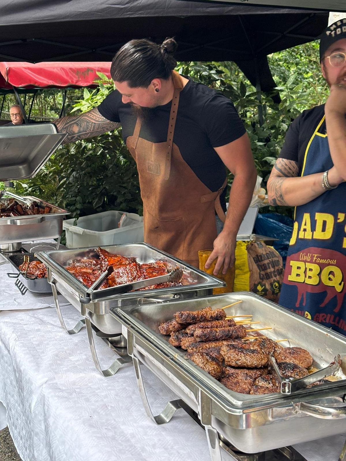 Man met tattoo's en een bruin apron serveert barbecue vlees op een buffet tafel, met twee andere mensen op een barbecue stand, omringd door groen tuin.