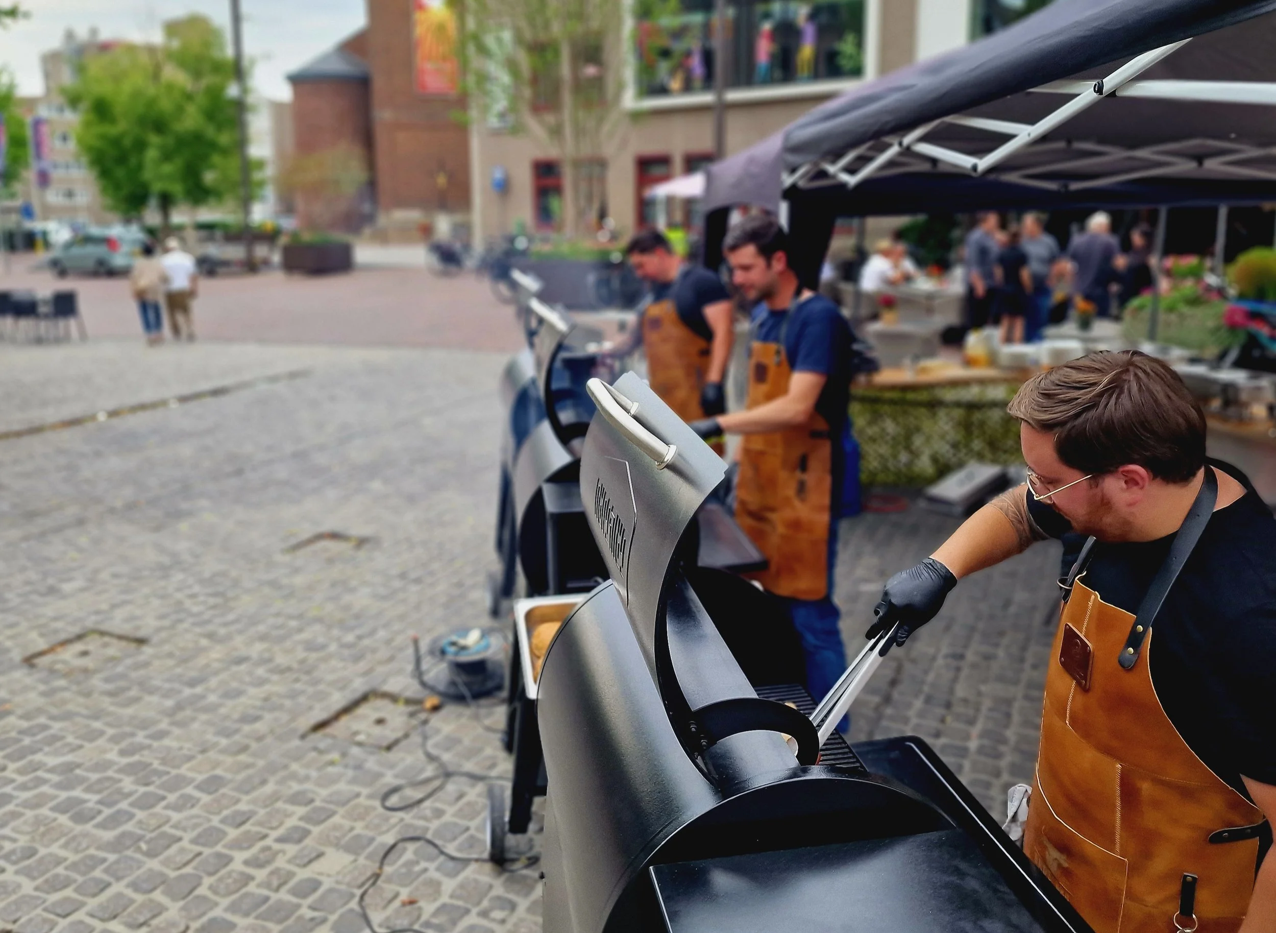 Mensen deden mee aan een foodmarkt, ze koken op barbecue met een tent op een plein. Ze dragen honingkleurige schorten.