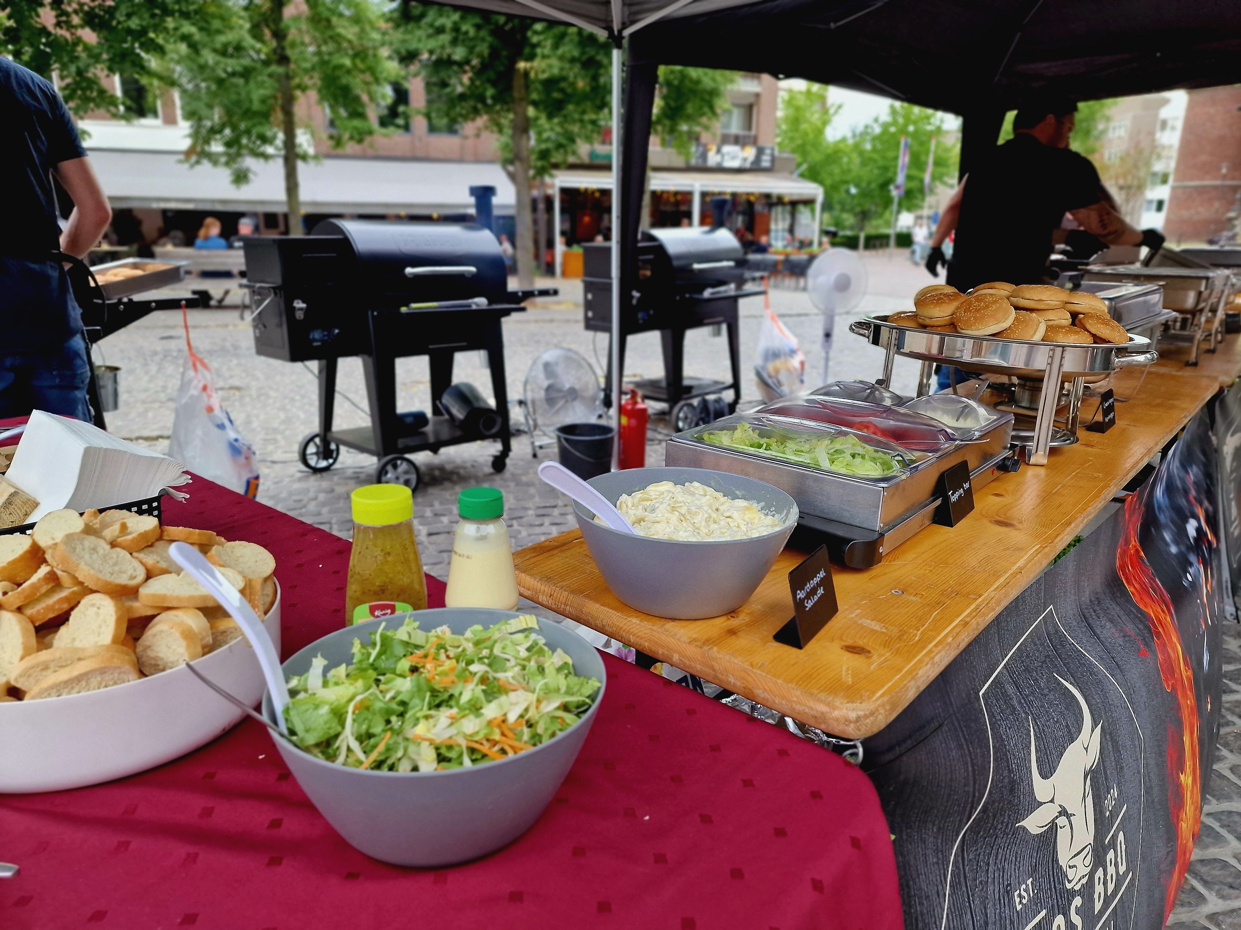 Buffet met broodjes, salade, saus, en toppings bij een food truck, met barbecue grills op de achtergrond.