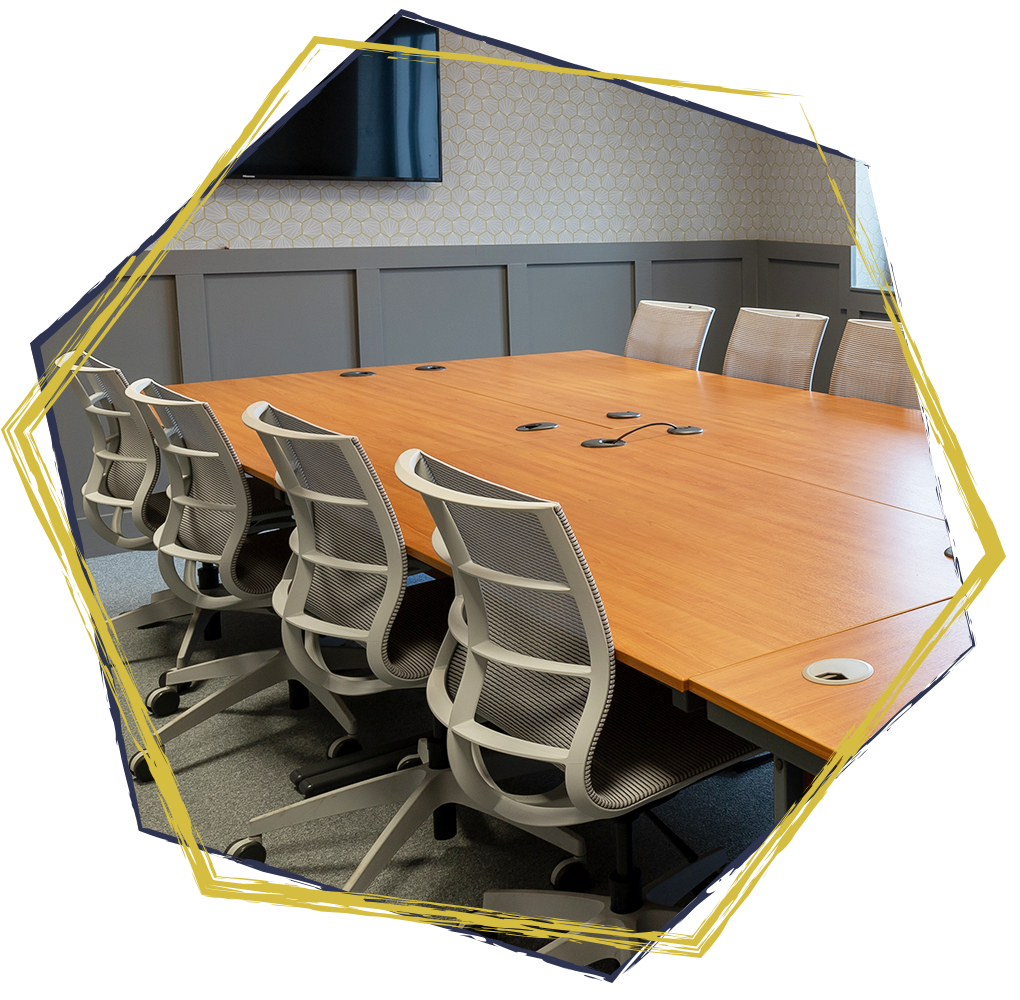 A conference room with a large wooden table and nine ergonomic office chairs with mesh backs, some featuring armrests. The room has a textured wallpaper with a pattern, gray wainscoting, and a window allowing natural light in. There are multiple built-in microphones on the table.