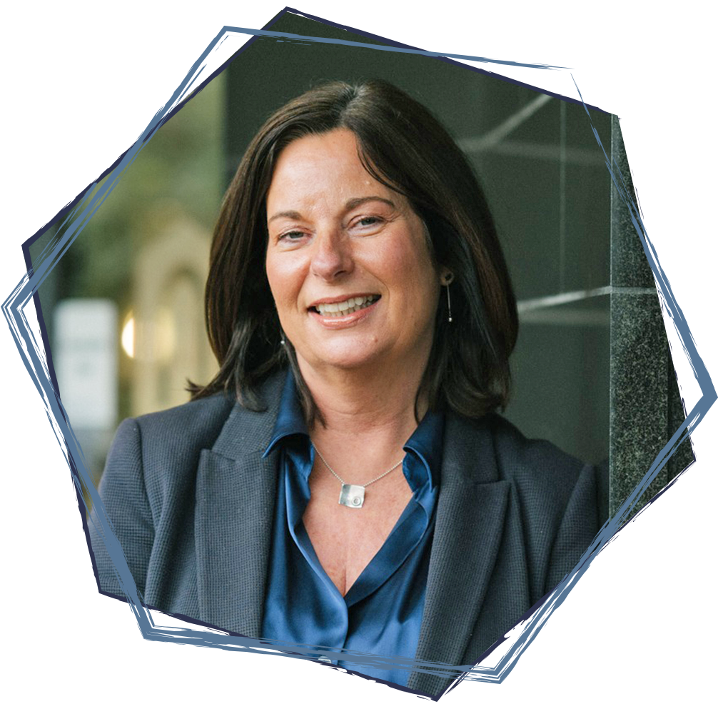 A middle-aged woman with dark brown hair smiling, wearing a dark gray blazer, a blue shirt, earrings, and a necklace, standing outdoors against a blurred background.