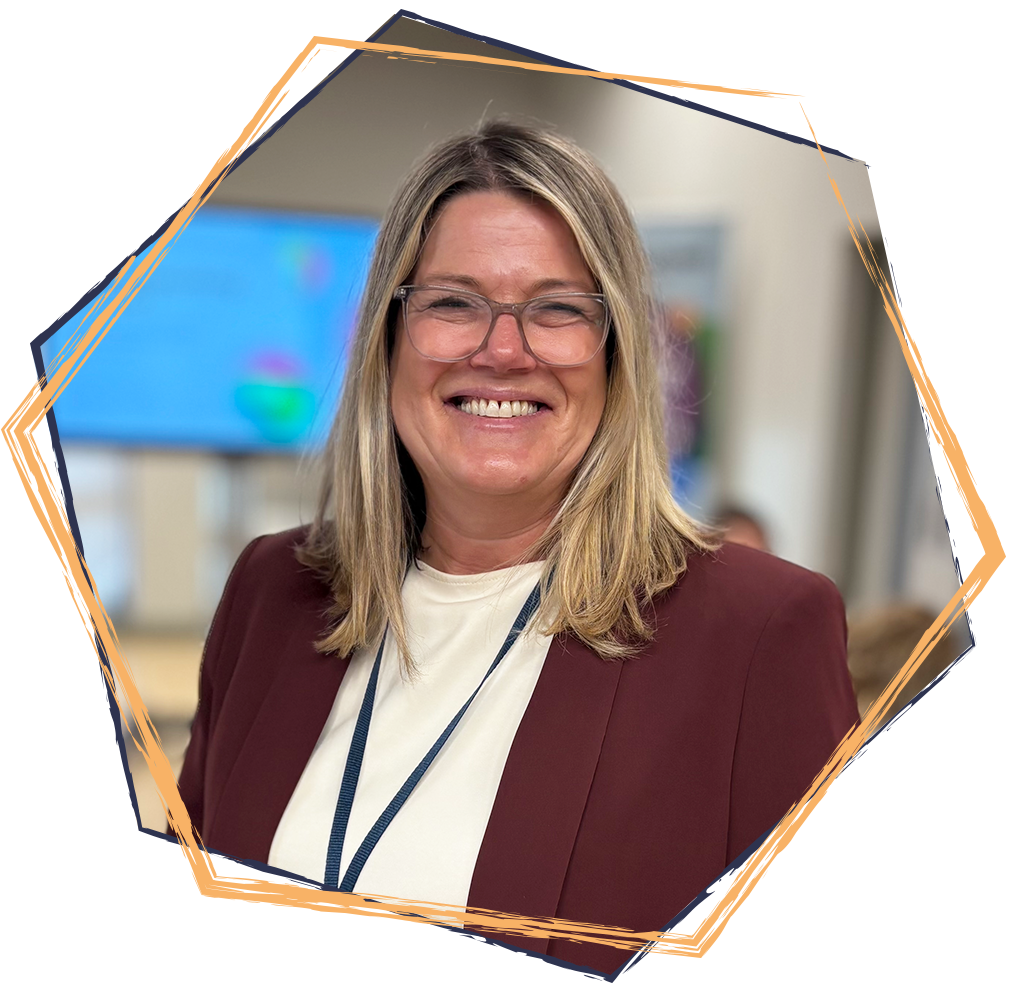 A smiling woman with blonde hair, glasses, and a maroon blazer standing indoors, possibly at a conference or event.