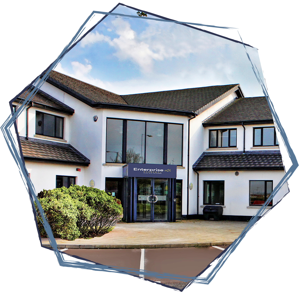 A white two-story building with black window frames and a glass entrance, labeled "Enterprise". Small bushes are in front, and a parking lot is partially visible. The sky is partly cloudy.