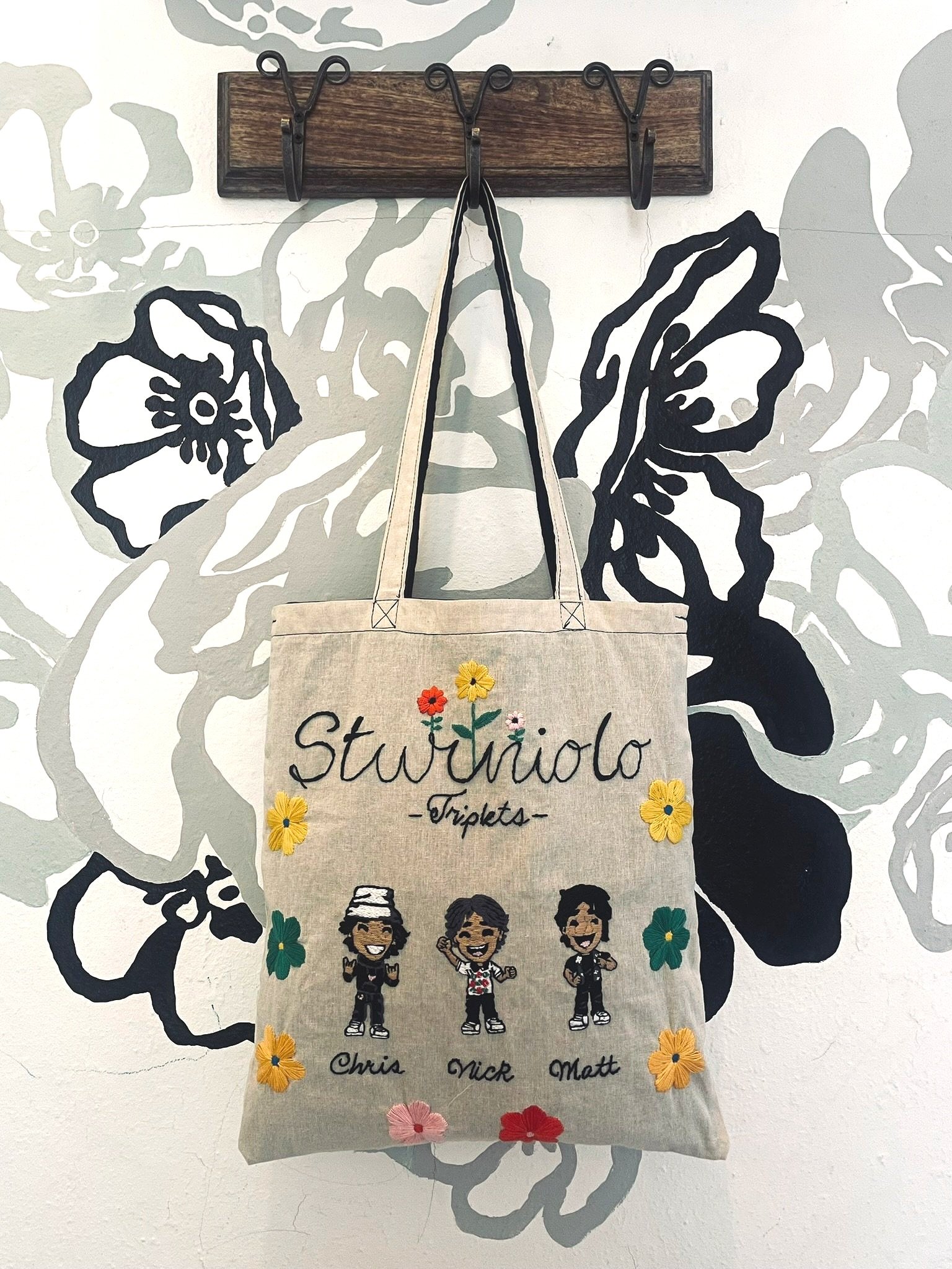 A tote bag featuring cartoon caricatures labeled Chris, Nick, and Matt, under the embroidered text 'Sturniolo Triplets,' surrounded by embroidered flowers. The bag hangs on a wall with a floral mural and wooden coat hooks.