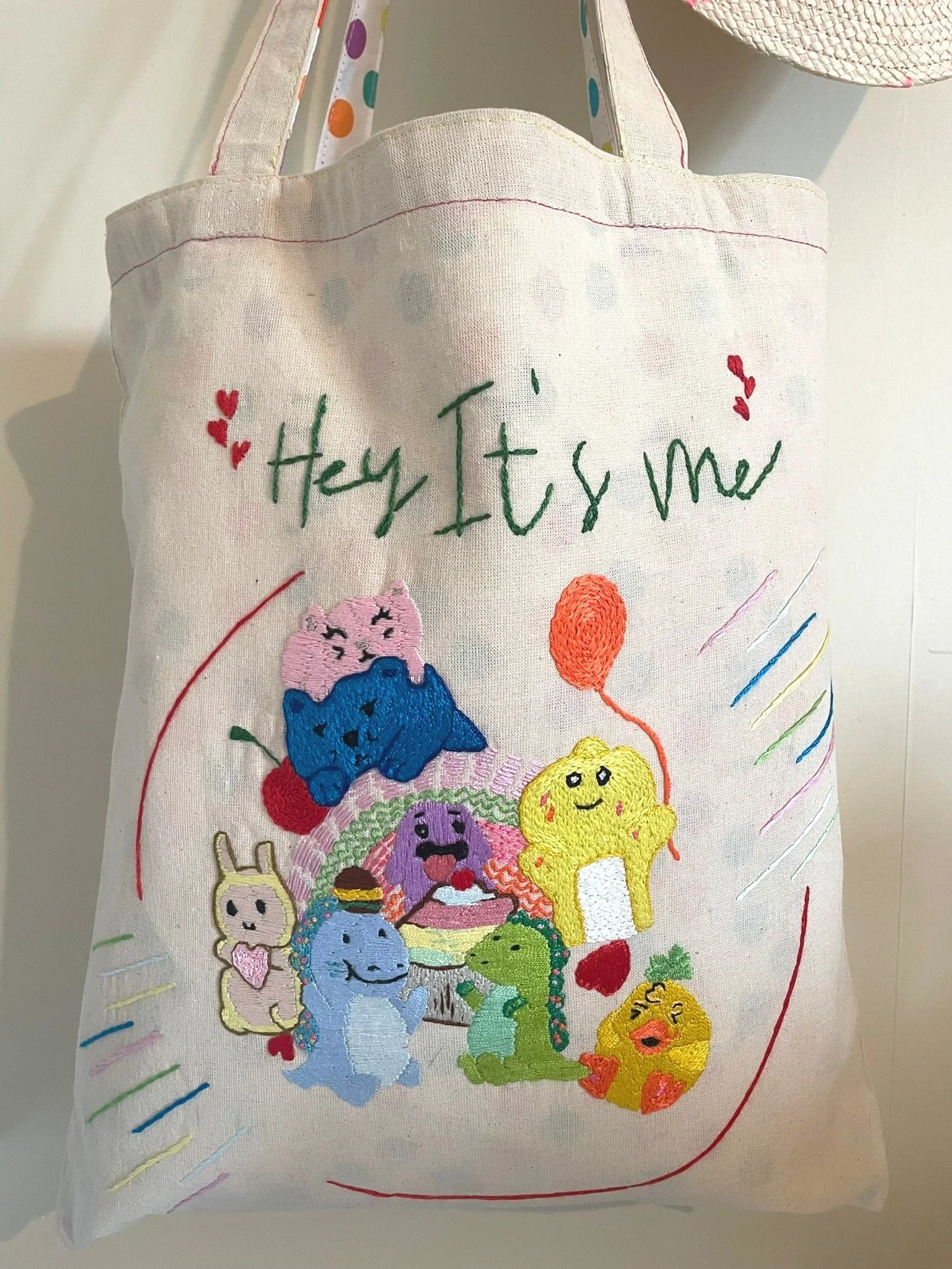 Embroidered tote bag with colorful cartoon animals, including a pink cat, blue bear, yellow character holding a balloon, and various creatures, surrounding a cake. Text reads 'Hey It's me' with heart motifs.