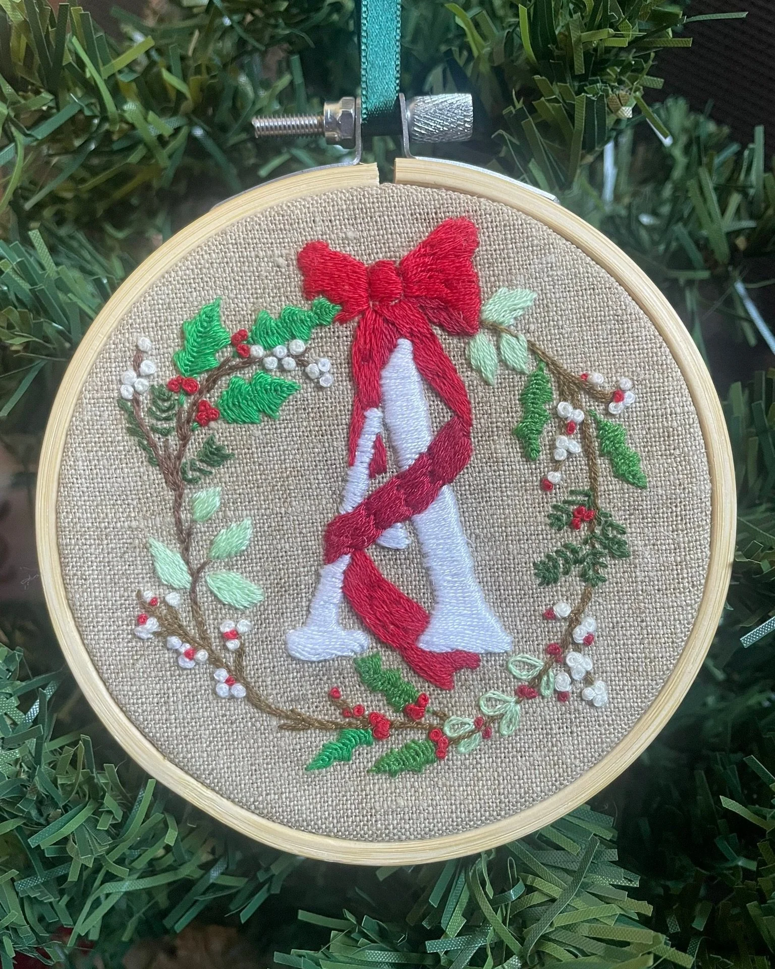 Embroidery hoop art with the letter 'A' wrapped in a red bow, surrounded by a wreath of embroidered holly and berries, hanging on a Christmas tree.