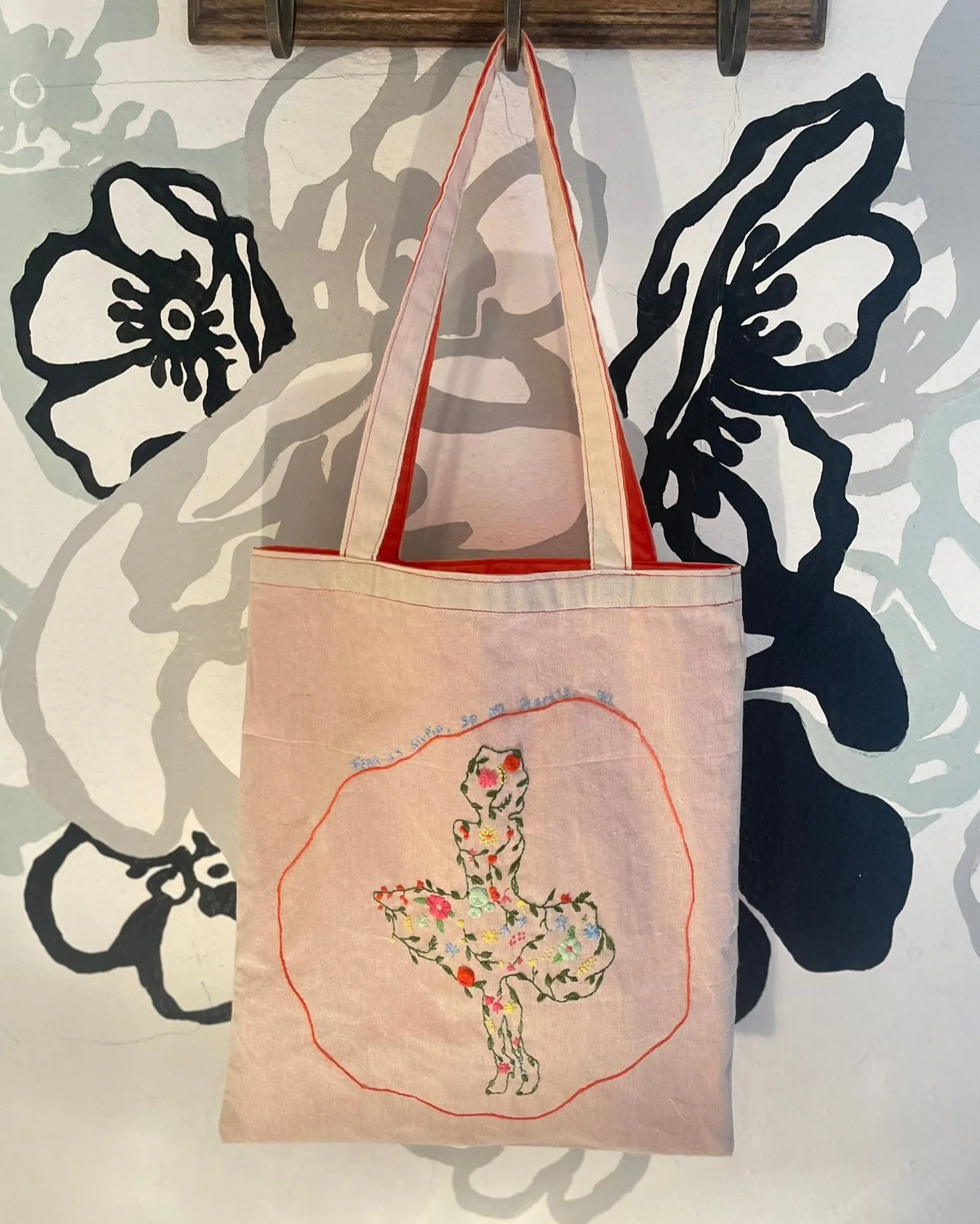Embroidered tote bag with the iconic 'Marilyn Monroe Silhouette' floral decoration hanging on a hook against a floral mural background.