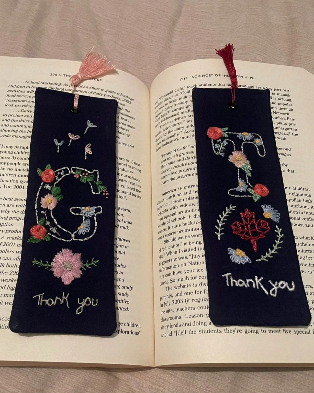 Two embroidered bookmarks with floral designs on a book; one with a 'G' and one with a 'T', each accompanied by the text 'Thank you', decorated with colorful flowers and tassels.