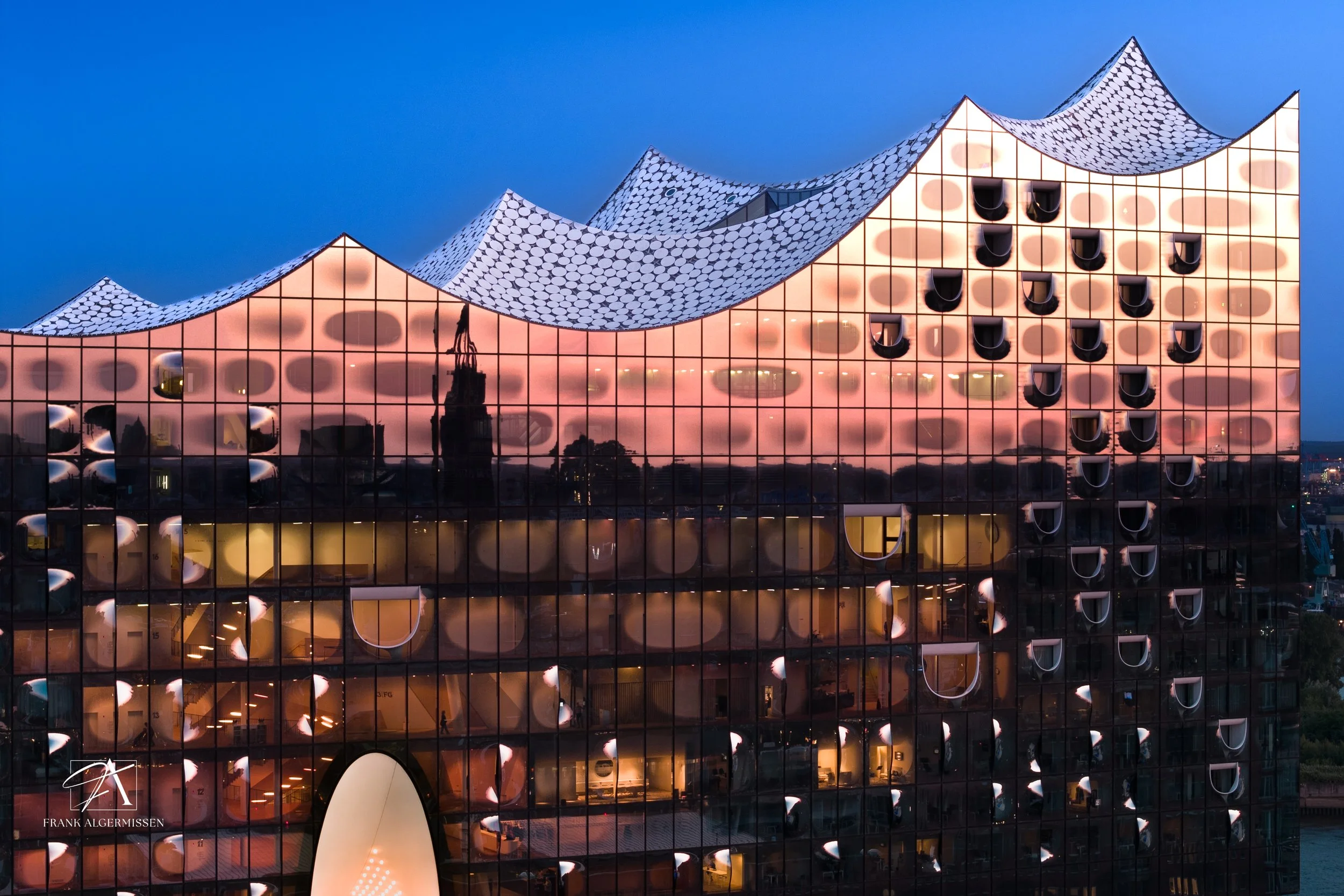 Modern building with reflective glass and a wavy, patterned roof, illuminated with soft pink and yellow lights at dusk.