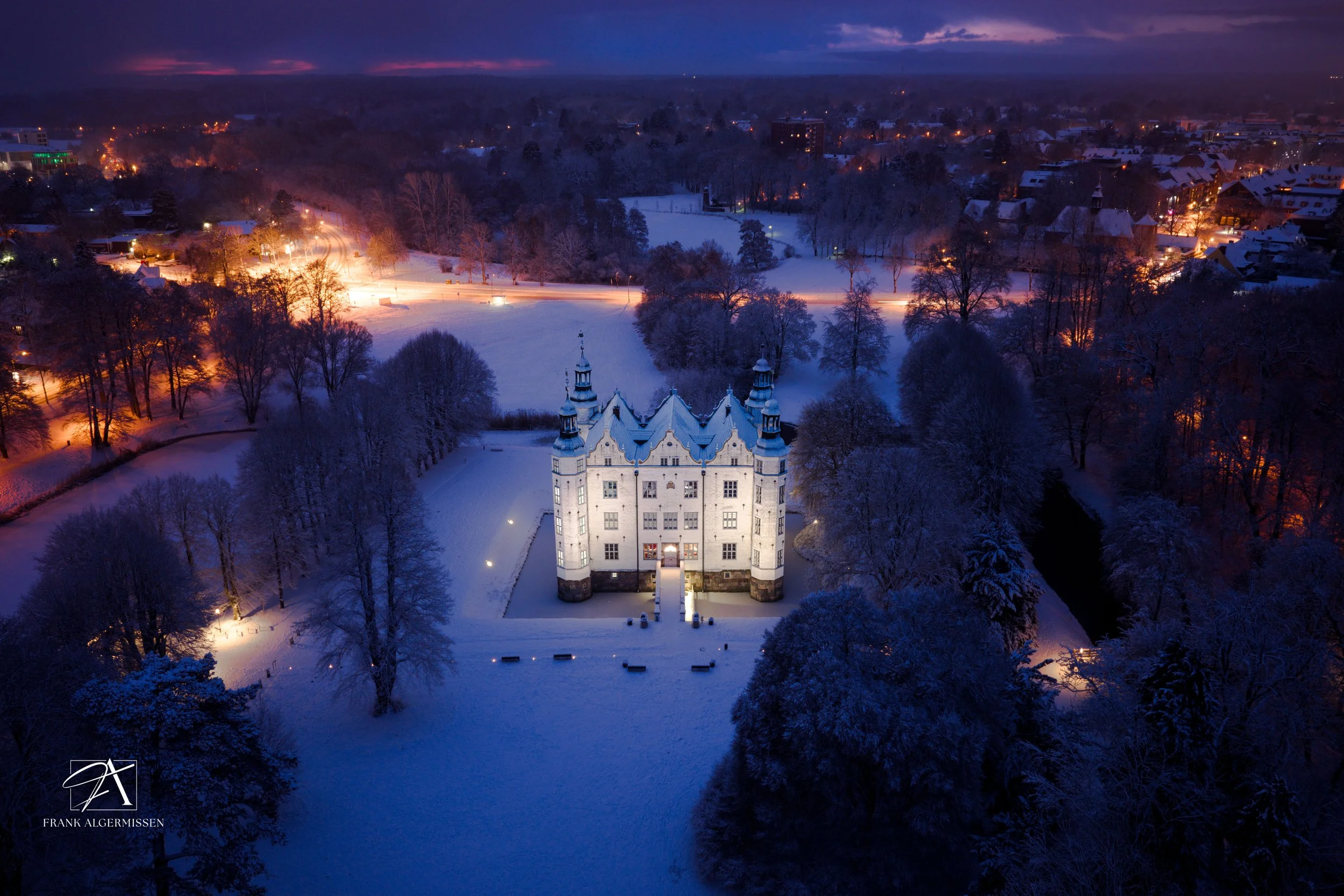 A snowy landscape with a illuminated Ahrensburg castle in the distance, surrounded by trees with snow-covered branches, under a golden purple morning sky