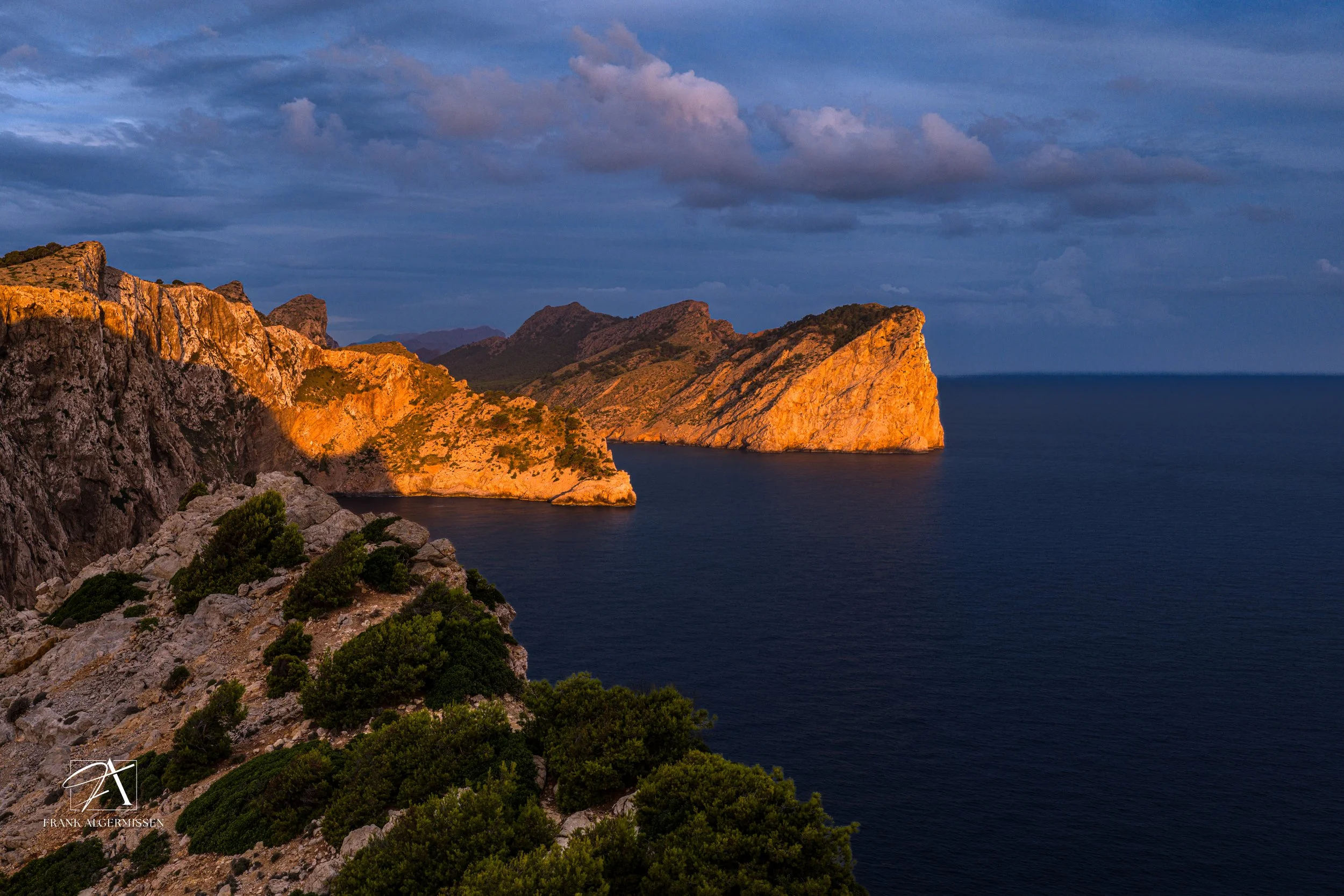 Sunlit coastal cliffs with green vegetation and calm dark blue sea, under a cloudy sky.