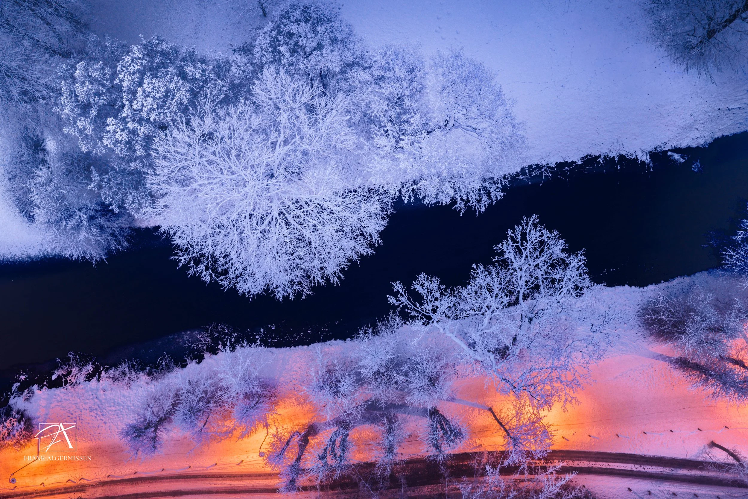 Bird's-eye view of snow-covered trees along a winding river at dusk, with a warm glow from streetlights illuminating the snow.