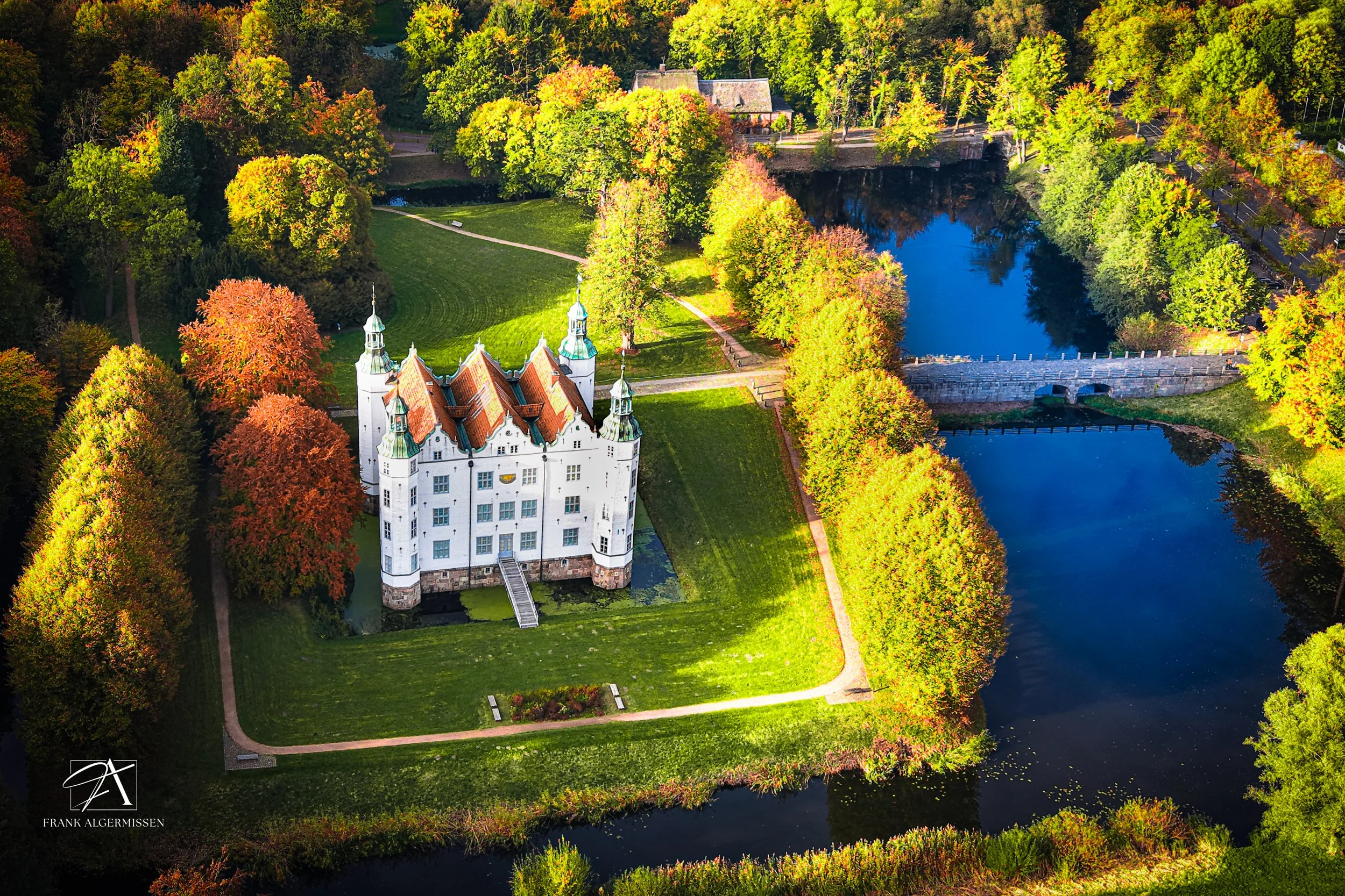 Aerial view of a fairy tale castle surrounded by a park with colorful autumn trees, green grass, and water features.