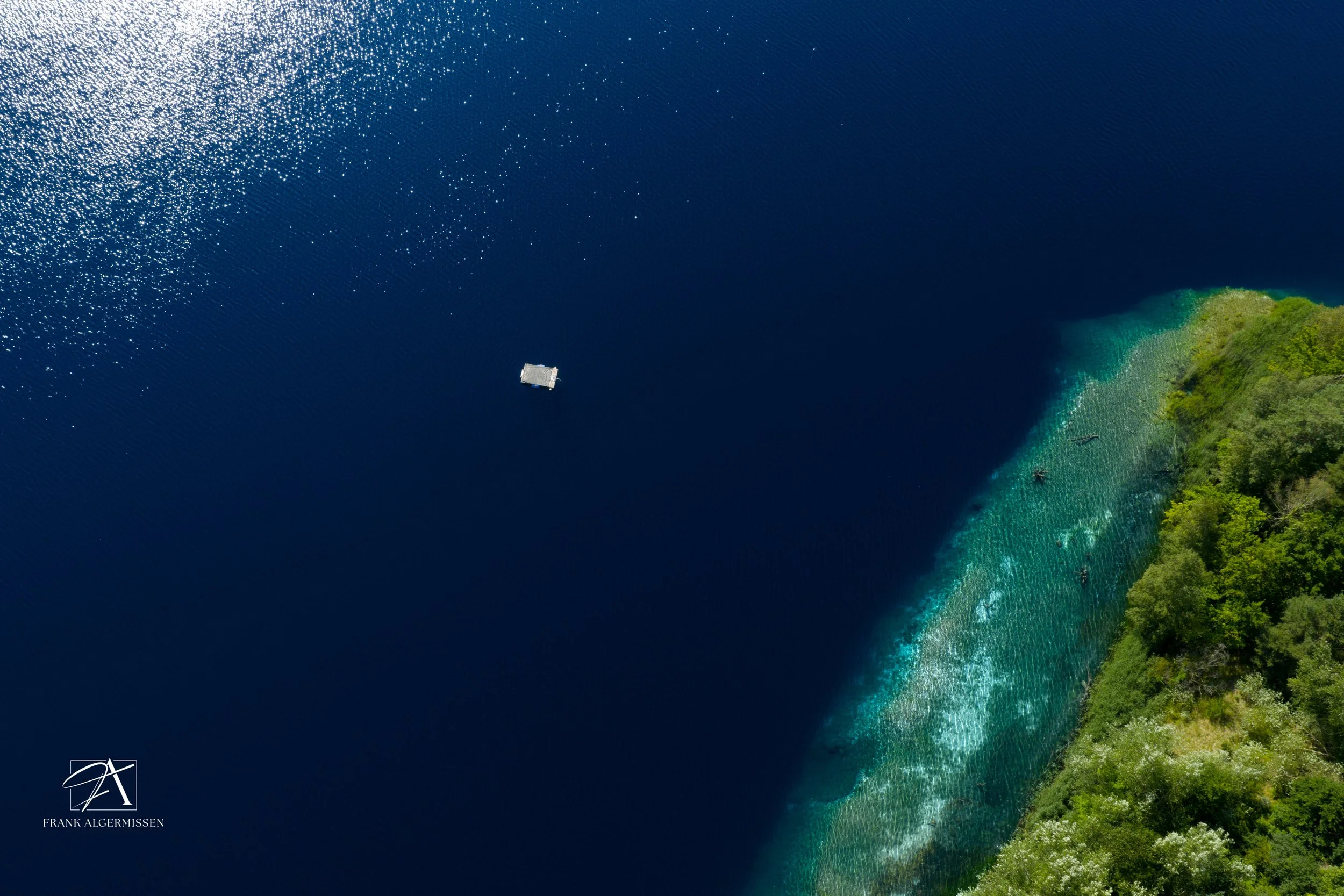 Aerial view of a swimming ponton on a deep blue lake with turquoise sandbanks and a green forested shoreline.