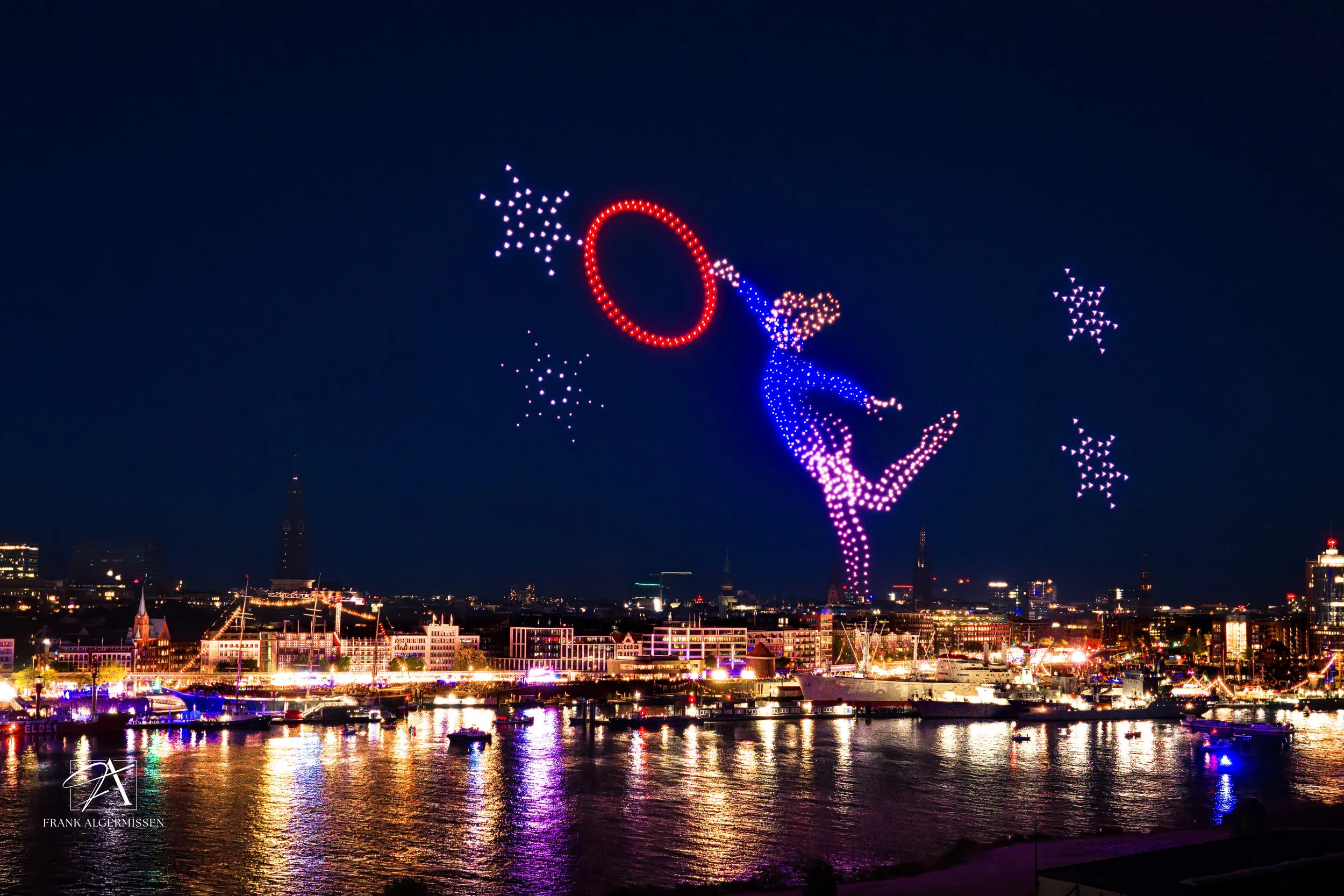 Nighttime cityscape with illuminated buildings, a harbor with boats and a large light display of a dancer with a red hoop and star-shaped lights in the sky created by 1000 drones for the 837. Hamburg Hafengeburtstag and its bid for the Olympia