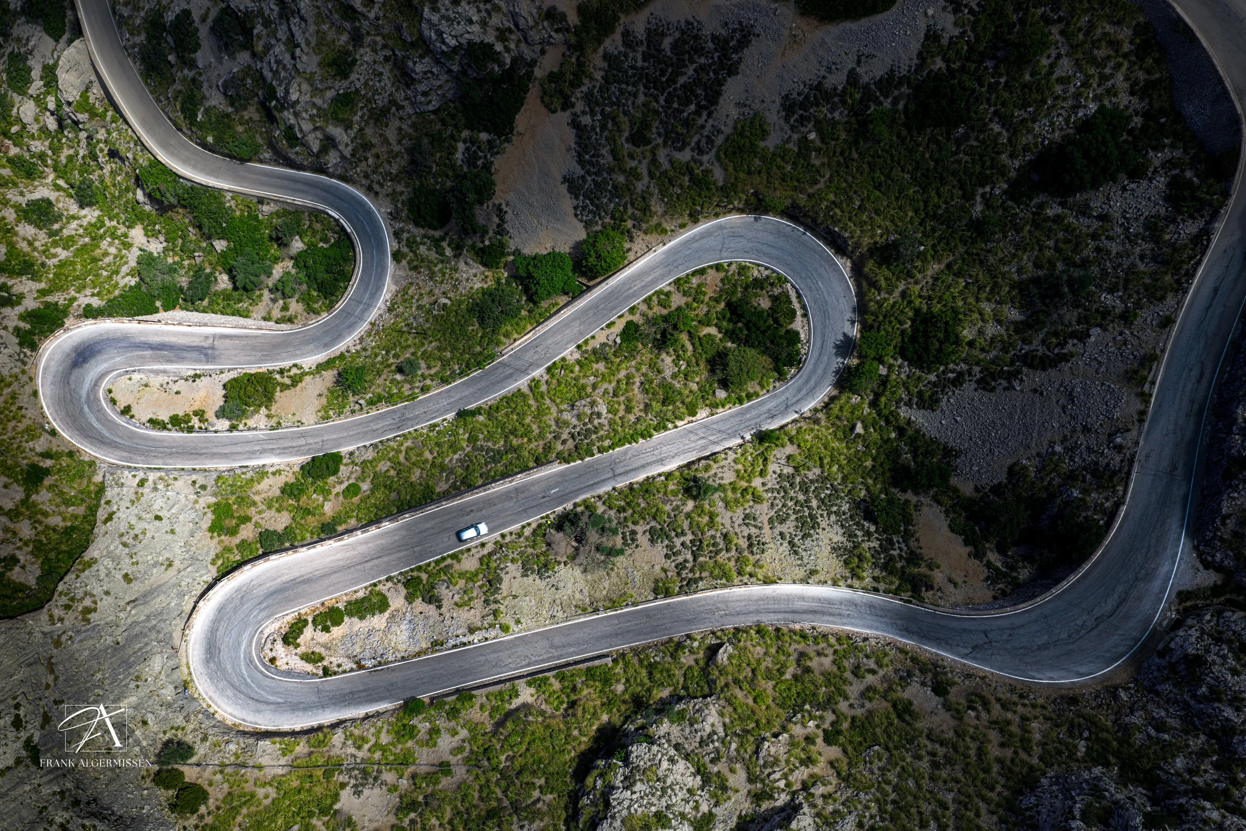 An aerial view of a winding mountain road with a single white car on it, surrounded by green and rocky terrain.