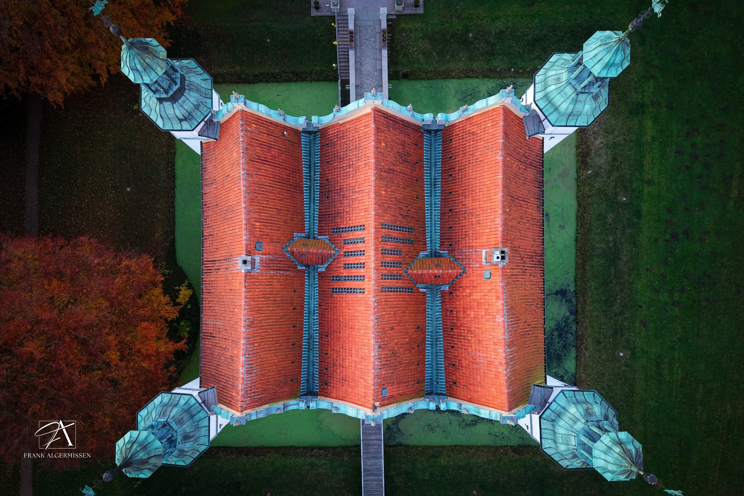 Aerial view of the Ahrensburg castle with a red tile roof and four towers, at sunset, surrounded by trees and grass.