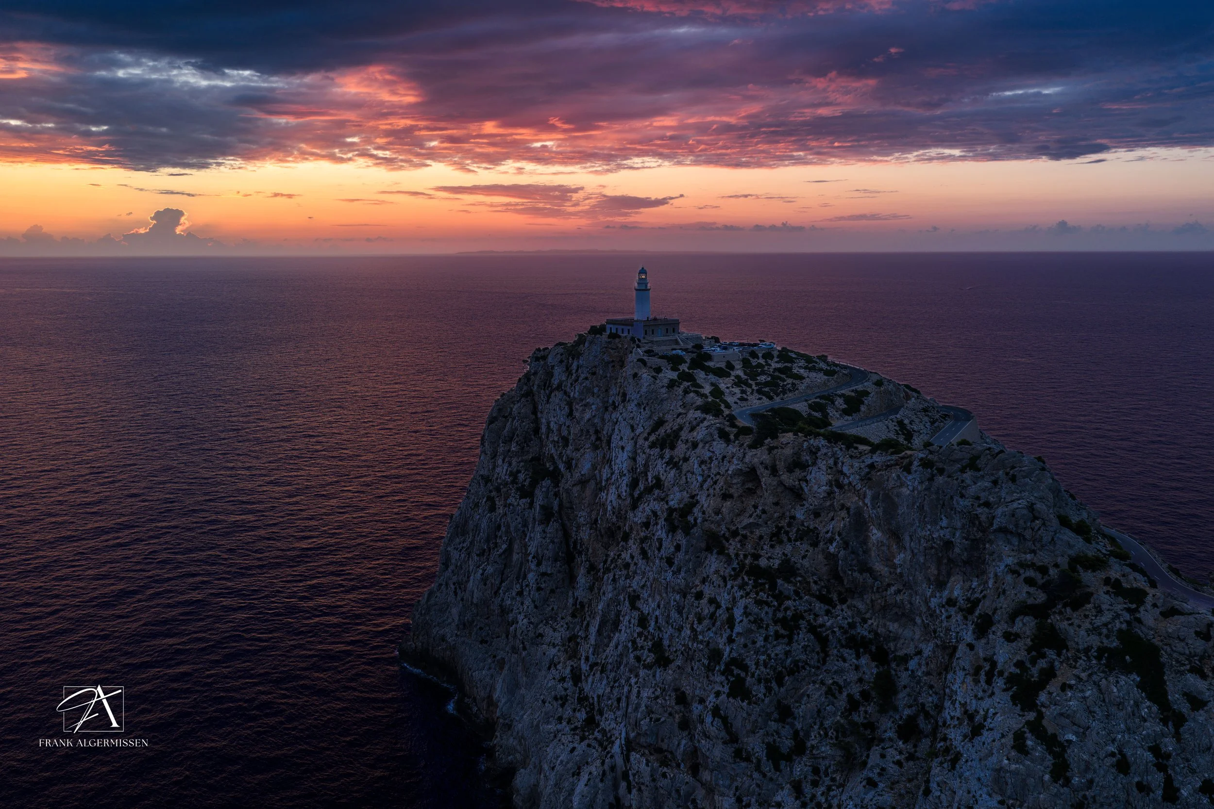 A lighthouse on top of a rocky cliff during sunset with a colorful sky and calm ocean in the background.