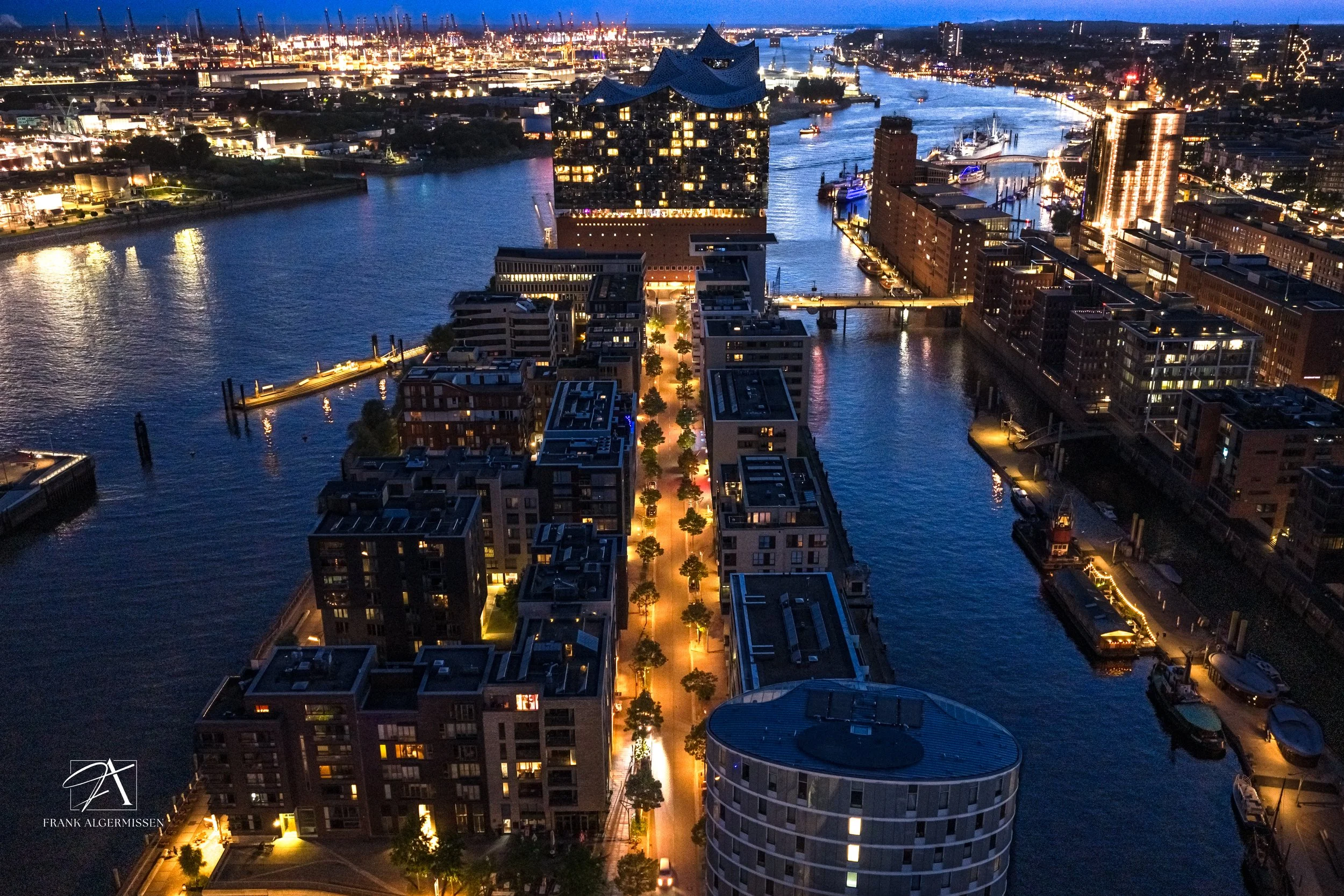 An aerial view of a city canal with modern buildings, illuminated streets, and a distinctive glass building at night.