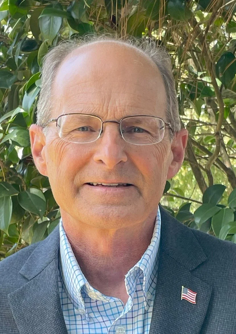 Close-up of an older man with glasses, wearing a checked shirt and a suit jacket, standing outdoors in front of green leafy foliage.
