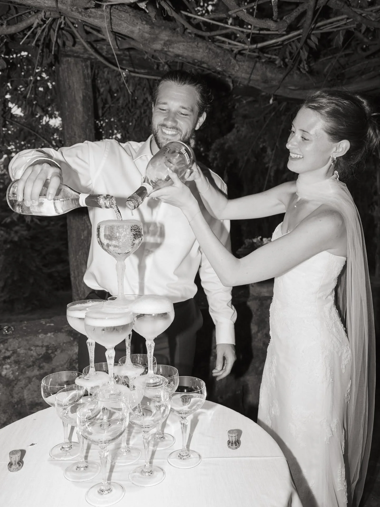 A bride and groom pouring champagne into a tower of glasses at their wedding reception in an outdoor setting.