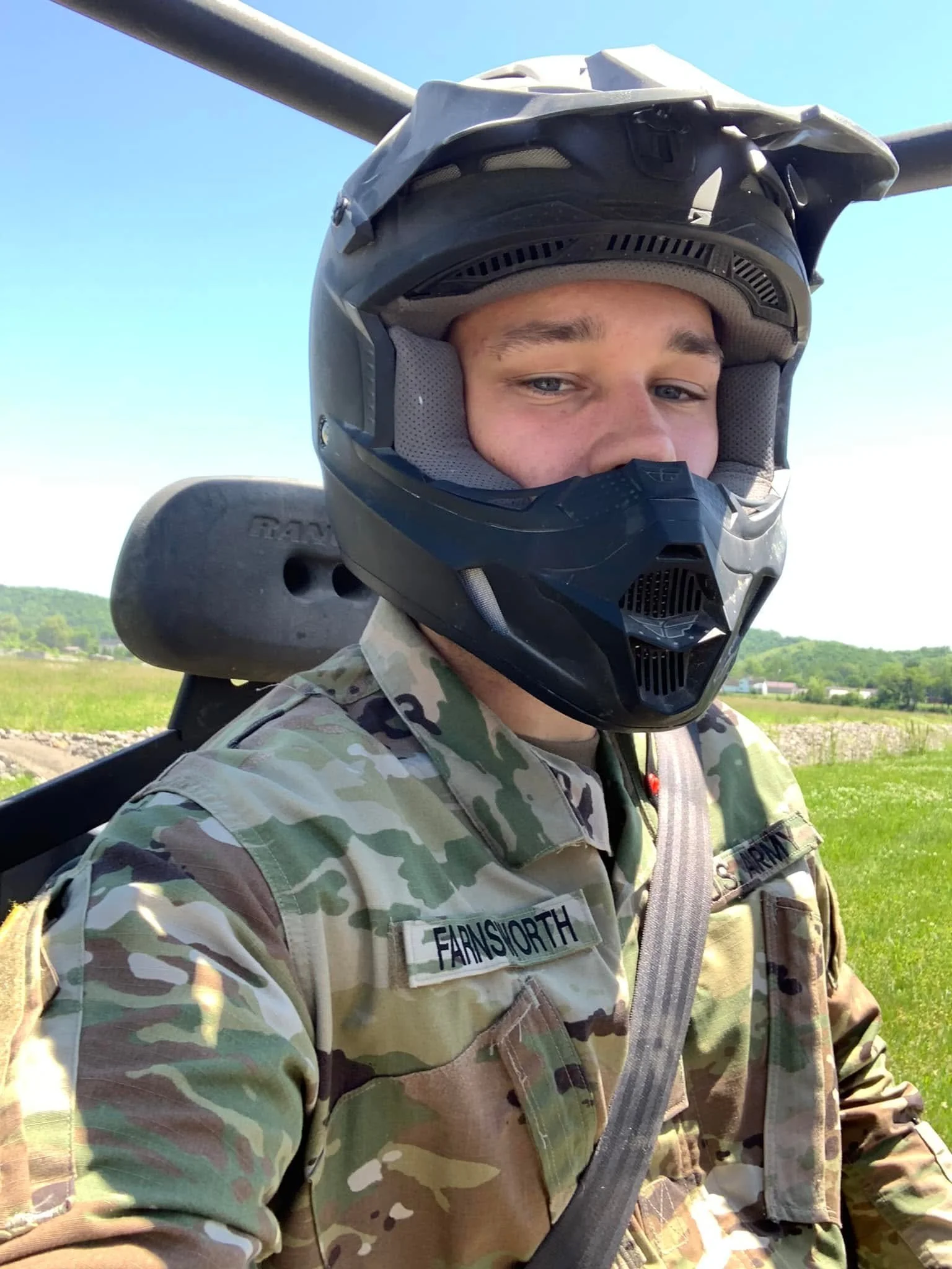 A person wearing a camouflage military uniform and a black helmet with a face shield, standing outdoors in a grassy area with blue sky and trees in the background.