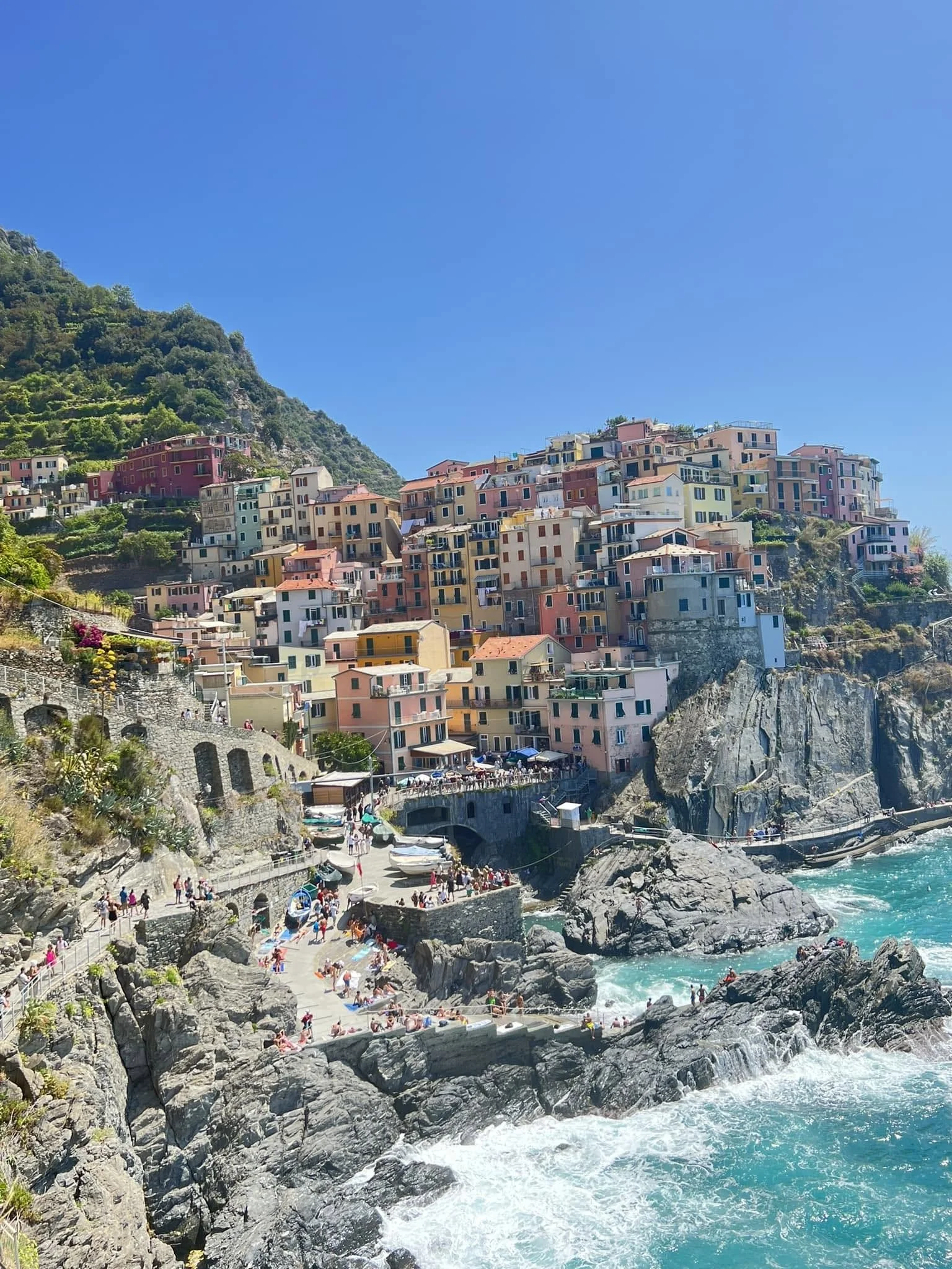 A scenic coastal town with colorful buildings on a hillside overlooking the ocean, with people on a rocky beach and waves crashing against the rocks, under a clear blue sky.
