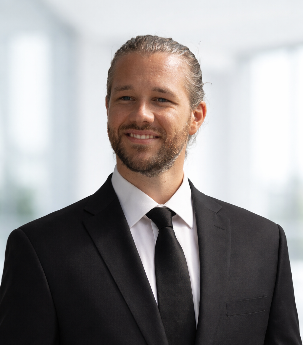 A man with long hair and a beard, wearing a black suit, white shirt, and black tie, smiling in an indoor professional setting.